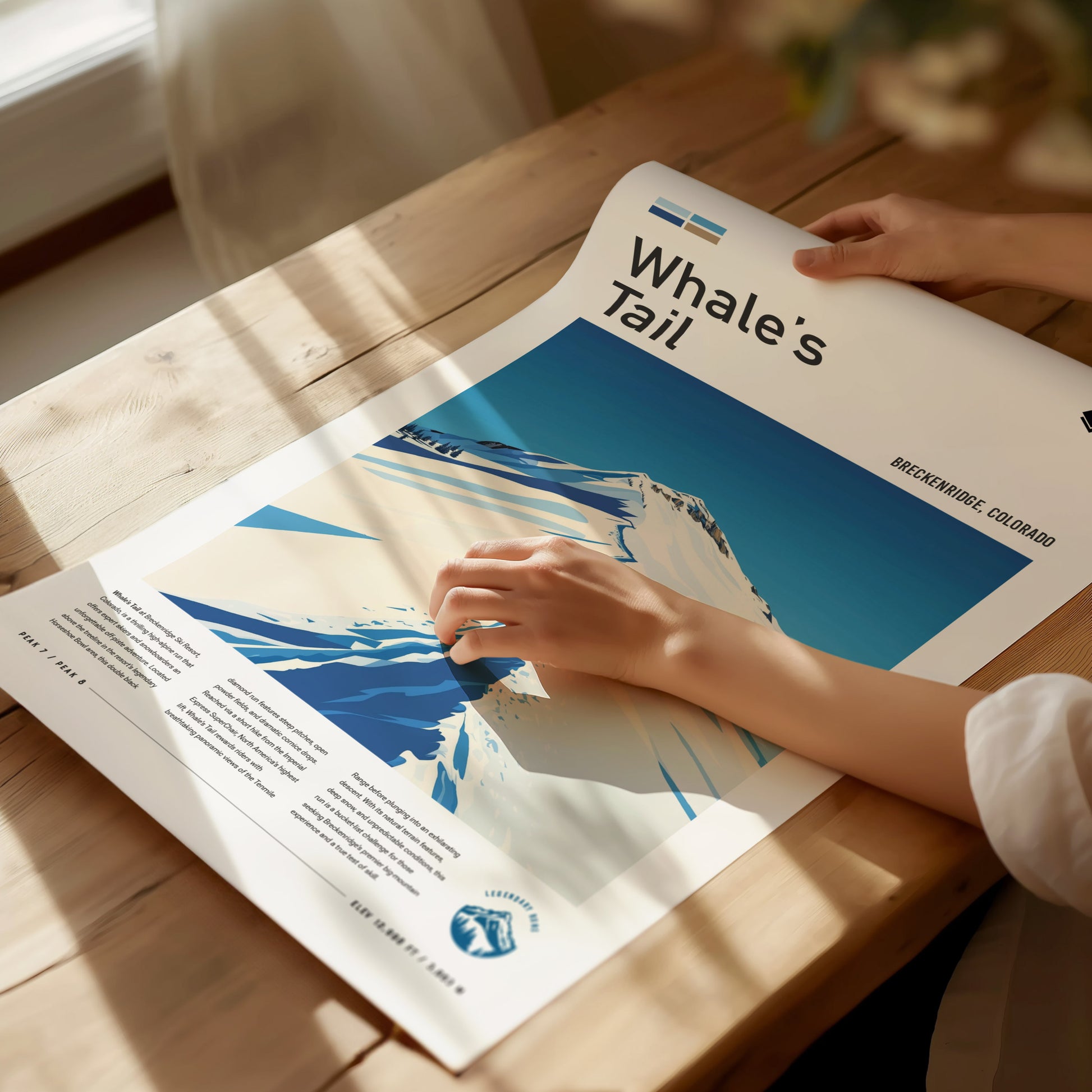 A person holds a Whales Tail Breckenridge poster on a wooden table, showcasing a stylized illustration of a snowy mountain ridge under a clear blue sky.