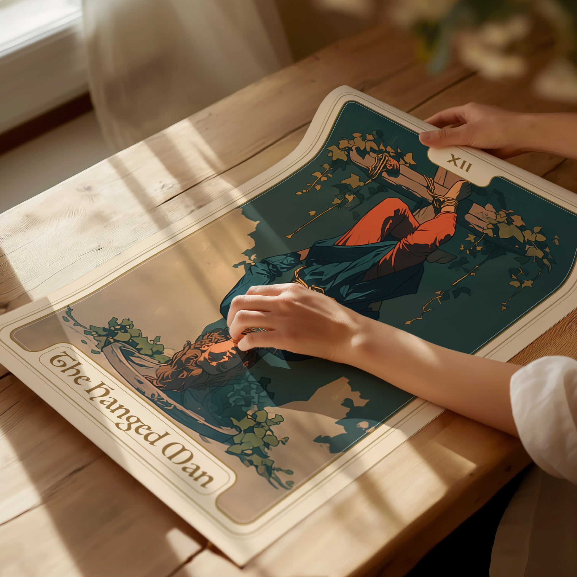 A person holds a Hanged Man Tarot poster on a wooden table, featuring an upside-down figure entwined with vines under sunlight. The card is labeled XII.