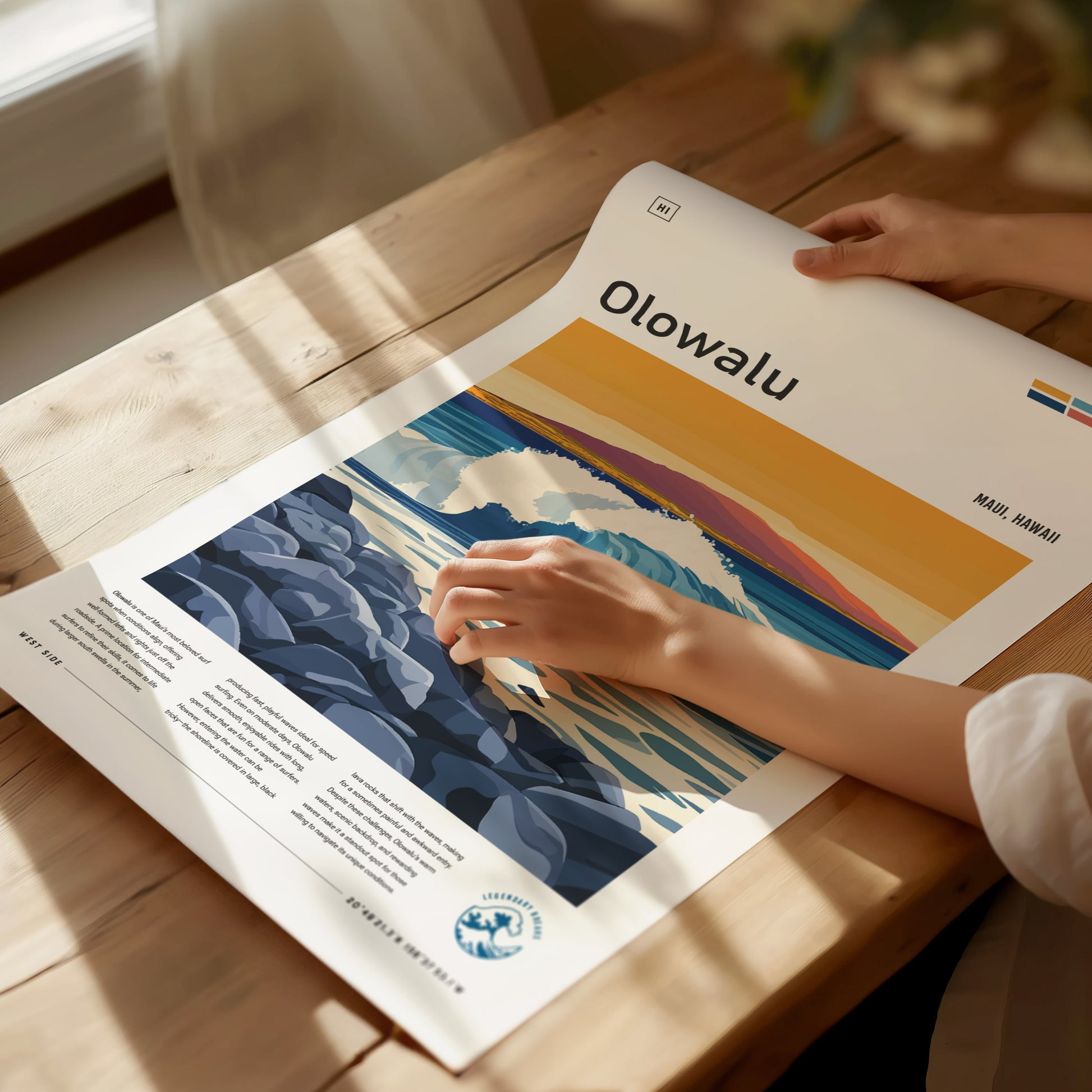 A person unrolls an Olowalu Maui surf poster on a wooden table, featuring vibrant colors of waves and rocky shoreline under a golden sky. Sunlight streams through a nearby window.