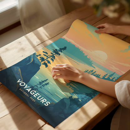 A person unrolls a colorful Voyageurs National Park poster on a wooden table, depicting a serene lake scene with trees and a sunset. Sunlight streams through a nearby window.
