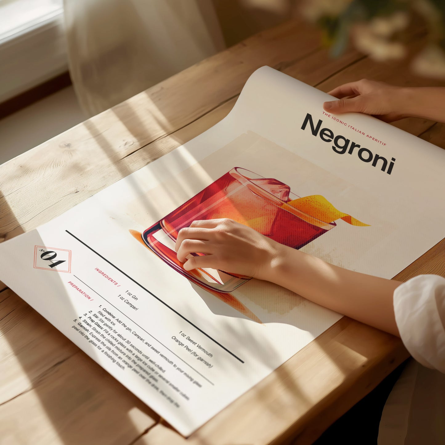 A person unrolls a Negroni cocktail poster on a wooden table, featuring an illustration of the drink with a citrus twist. Sunlight casts soft shadows across the scene.