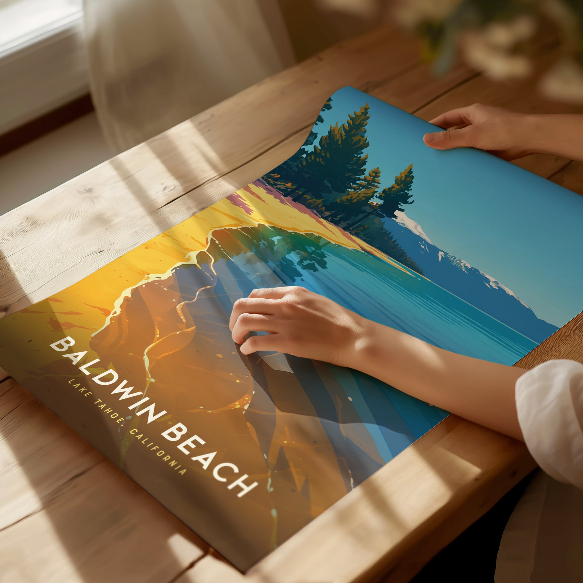 A person arranges a colorful Baldwin Beach, Lake Tahoe poster on a wooden table, showcasing vibrant waters, sandy shores, and evergreen trees under a clear blue sky.