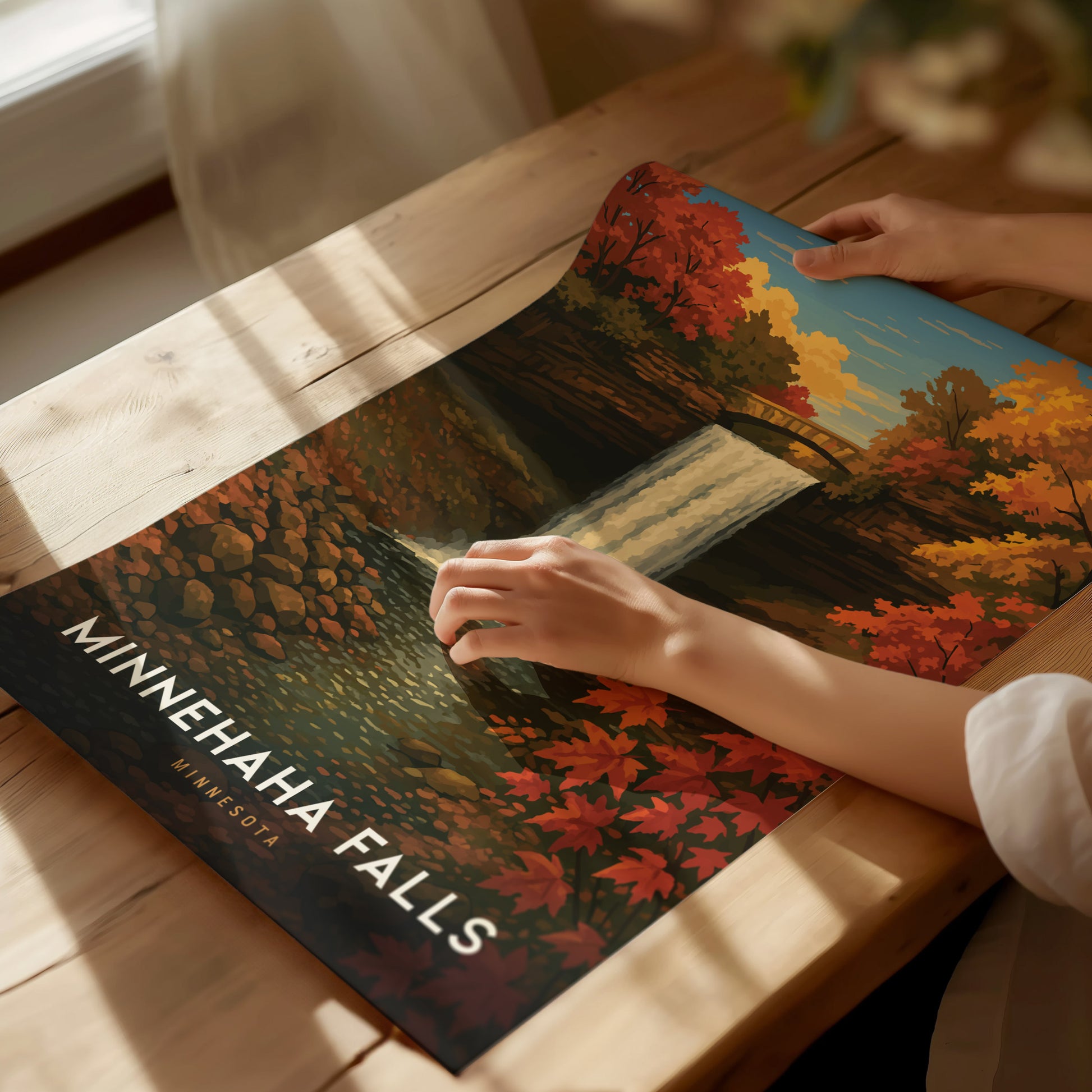 A person adjusts a Minnehaha Falls poster on a wooden table, featuring a waterfall and vibrant autumn foliage under a clear sky. Sunlight softly illuminates the scene.
