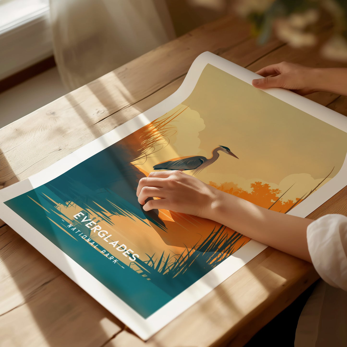 A person holds a minimalist Everglades poster featuring a heron by the water under a sunset. The art print is placed on a wooden table with soft lighting.