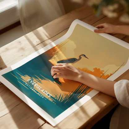 A person holds a minimalist Everglades poster featuring a heron by the water under a sunset. The art print is placed on a wooden table with soft lighting.
