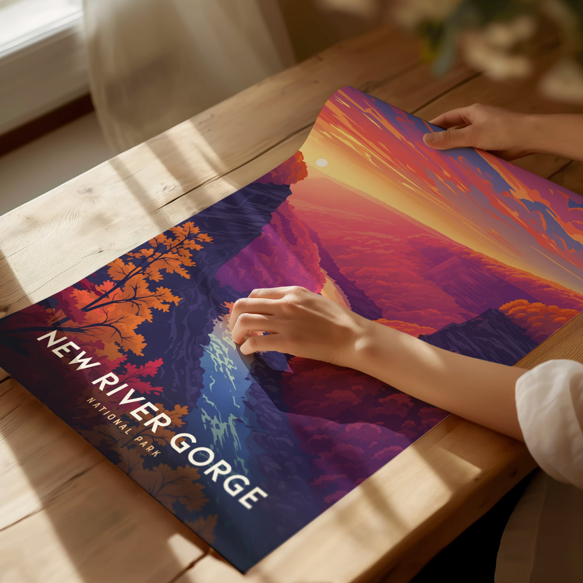A person adjusts a vibrant New River Gorge National Park poster on a wooden table, showcasing a colorful landscape with a sunset, rolling hills, and a river.