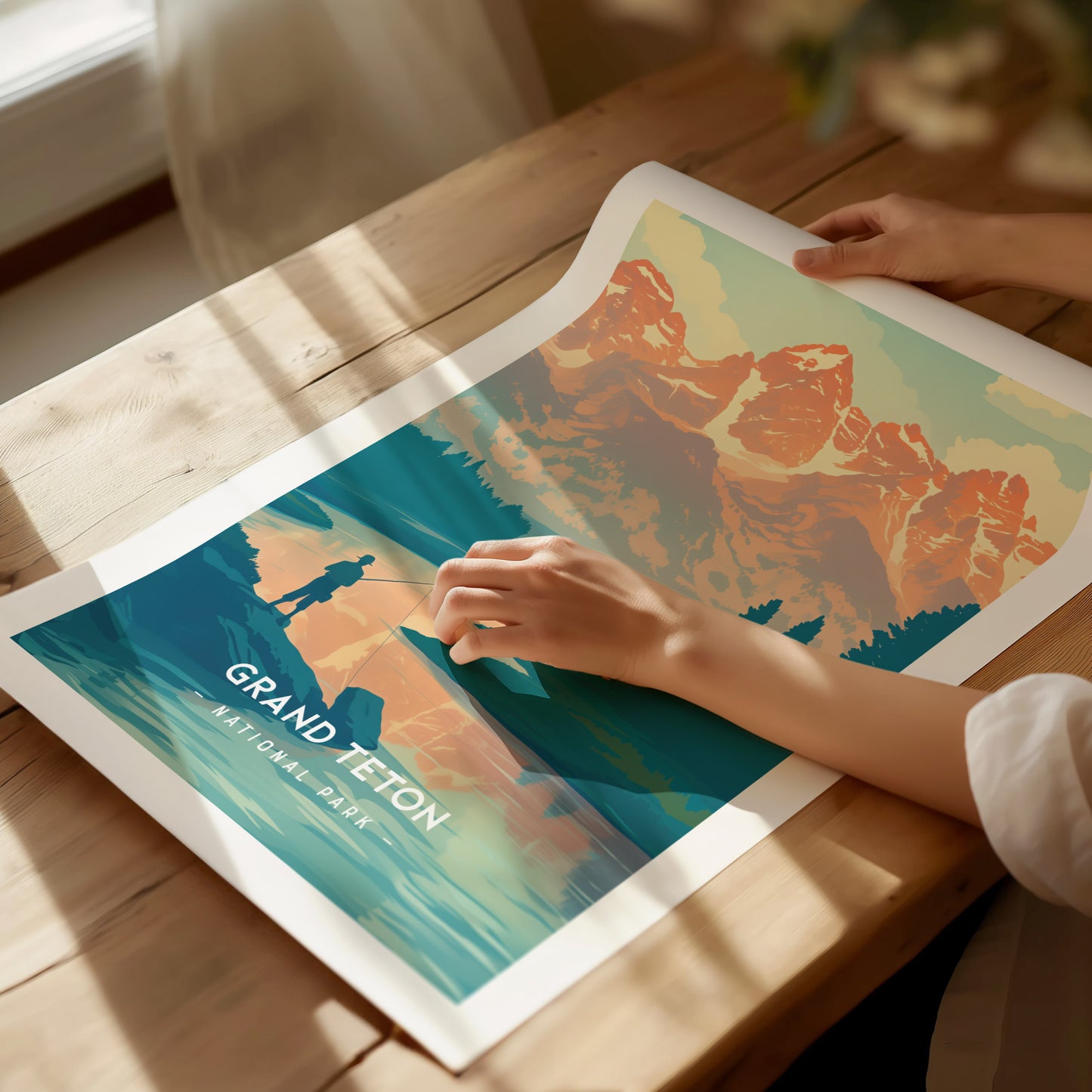 A person holds a vibrant Grand Teton National Park poster featuring a minimalist design with mountains and a fisherman. Sunlight casts shadows on a wooden table beneath the poster.