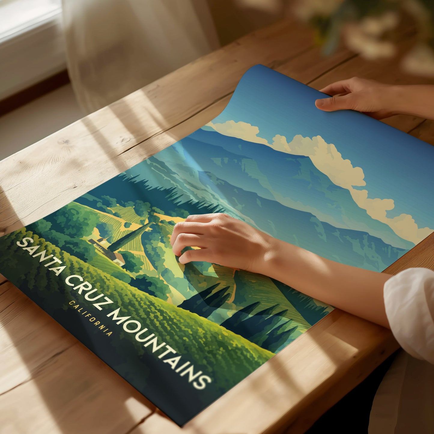 A person unrolls a colorful poster of the Santa Cruz Mountains on a wooden table, depicting lush green vineyards and majestic mountain peaks under a blue sky.