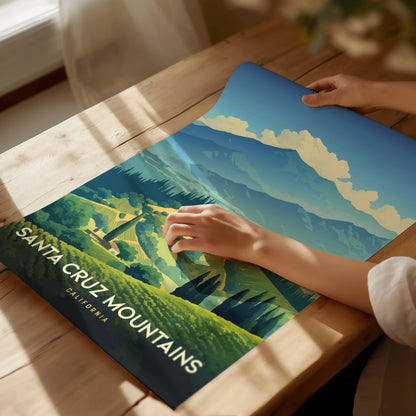 A person unrolls a colorful poster of the Santa Cruz Mountains on a wooden table, depicting lush green vineyards and majestic mountain peaks under a blue sky.