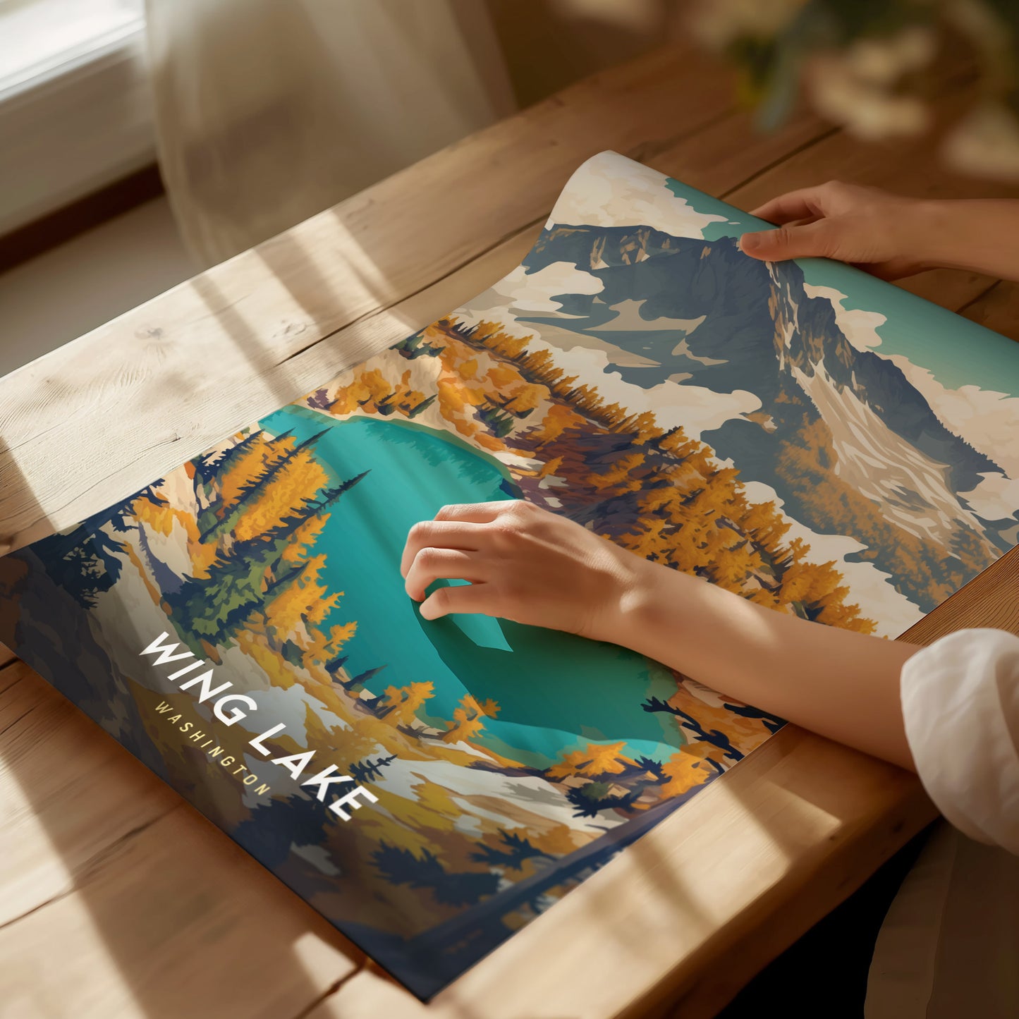 A person unrolls a colorful Wing Lake Washington poster on a wooden table, depicting vibrant autumn trees and a turquoise lake surrounded by mountains under a clear sky.