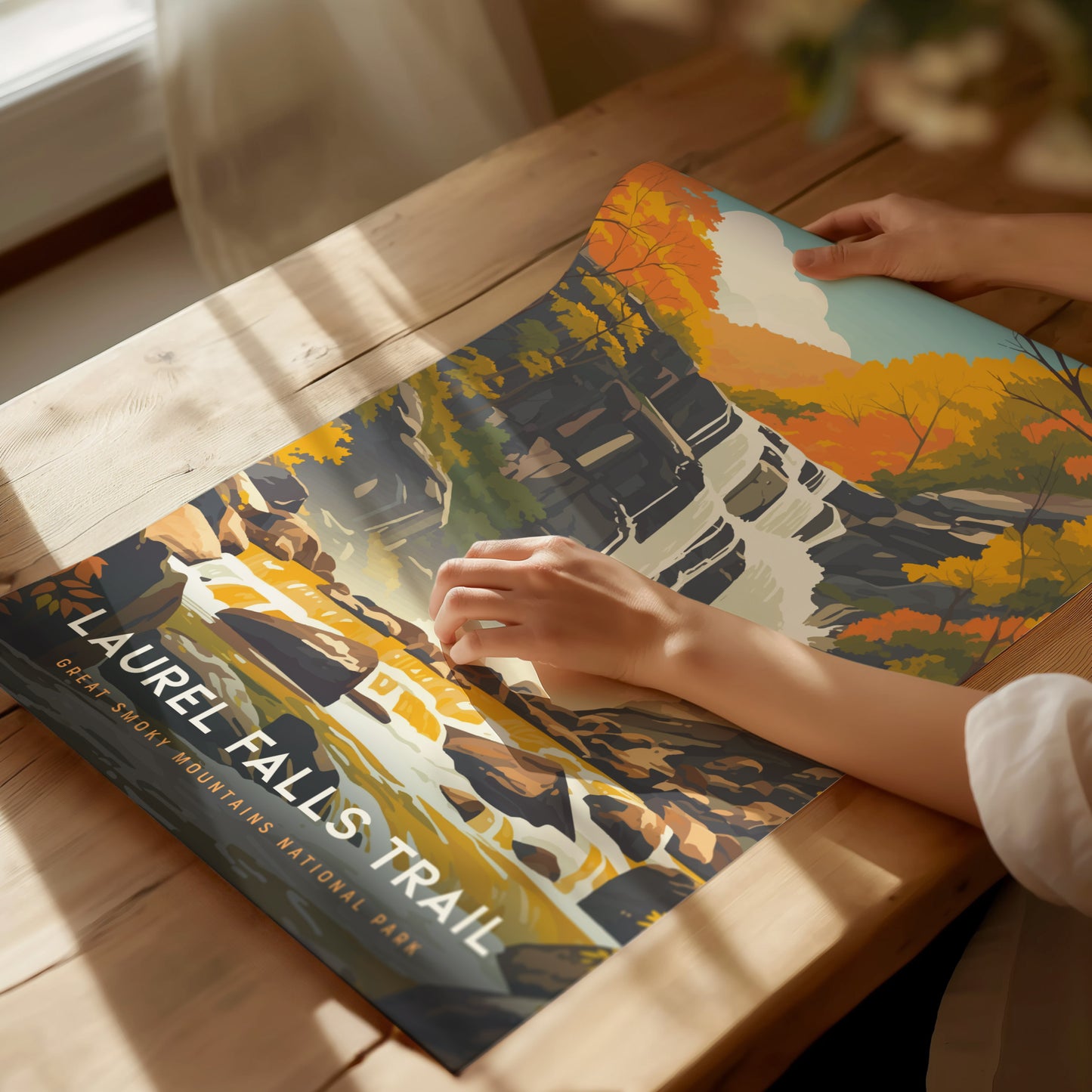 A person holds a colorful poster of Laurel Falls Trail featuring a waterfall and autumn foliage, on a wooden table with soft natural light.