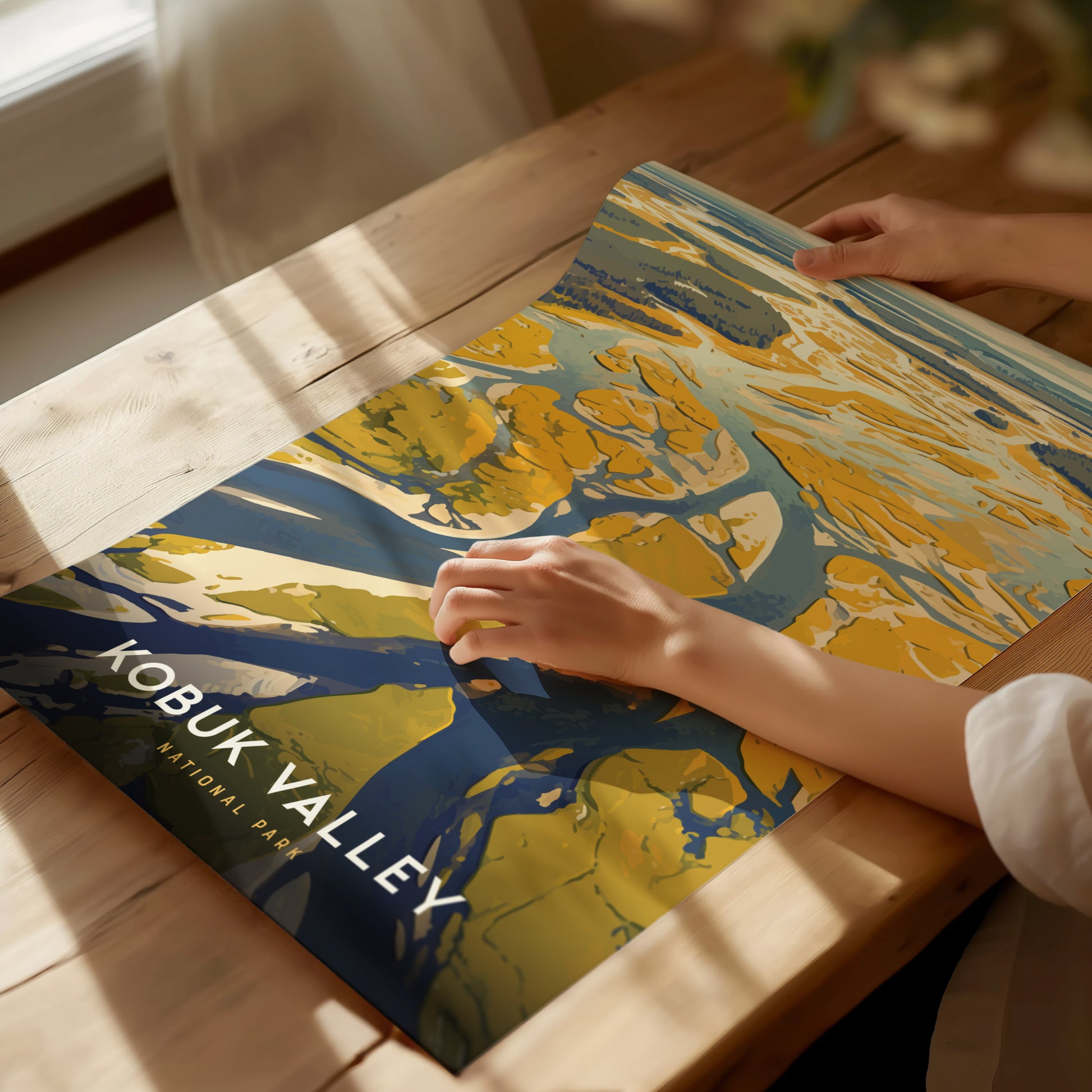 A person holds a colorful Kobuk Valley National Park poster featuring abstract sand dunes and rivers on a wooden table, with soft natural light illuminating the scene.