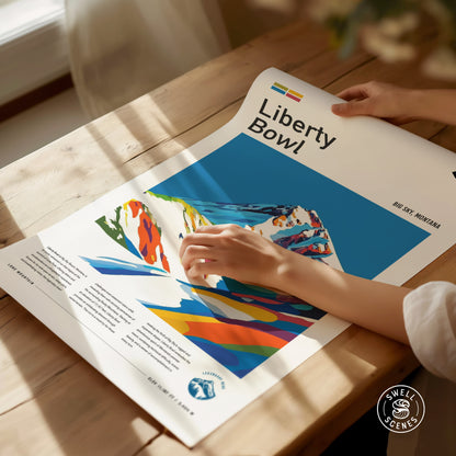 A person gently touches a colorful Liberty Bowl poster on a wooden table, featuring vibrant mountain artwork and text that reads Big Sky, Montana. Sunlight casts soft shadows.