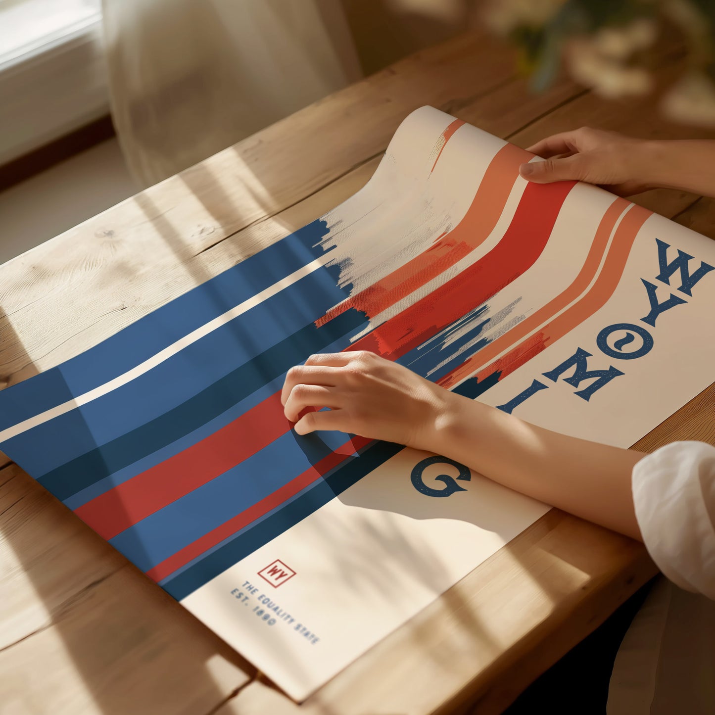 A person holds a colorful poster with bold red, white, and blue geometric shapes and the word WYOMING in stylized letters on a wooden table.