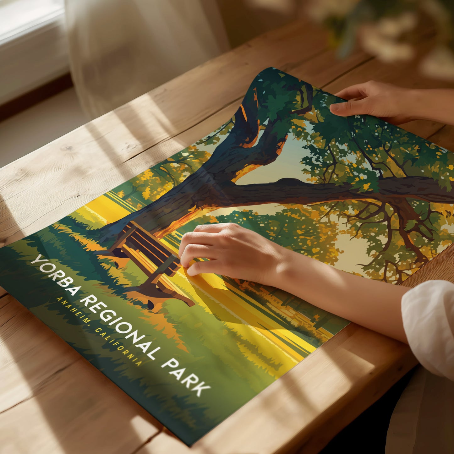 A person holds a Yorba Regional Park poster on a wooden table, depicting a serene landscape with a large tree and a bench. Sunlight softly illuminates the scene.