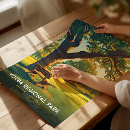 A person holds a Yorba Regional Park poster on a wooden table, depicting a serene landscape with a large tree and a bench. Sunlight softly illuminates the scene.
