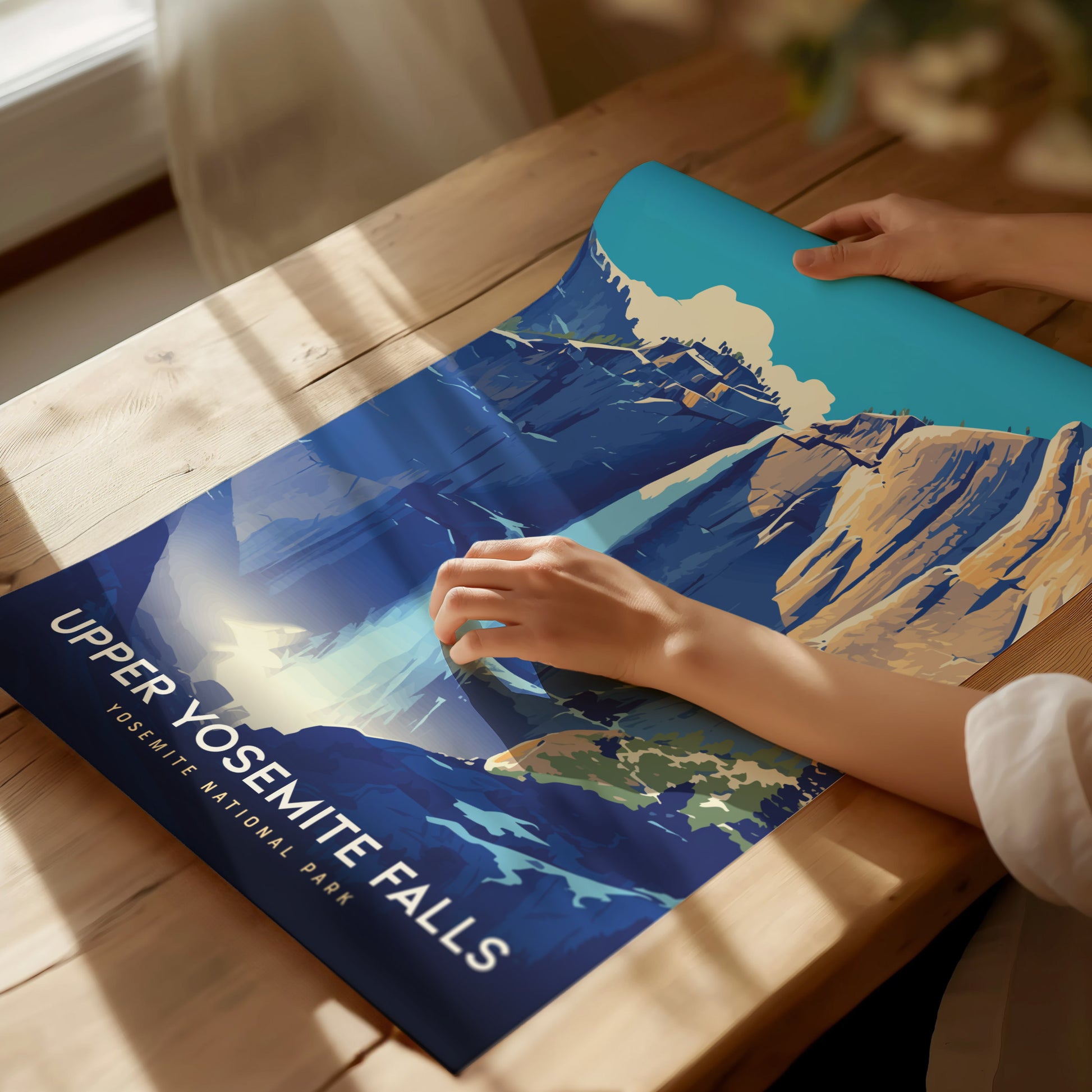 A person holds a colorful poster of Upper Yosemite Falls on a wooden table, with sunlight casting soft shadows. The artwork depicts the iconic waterfall against a vibrant blue sky.