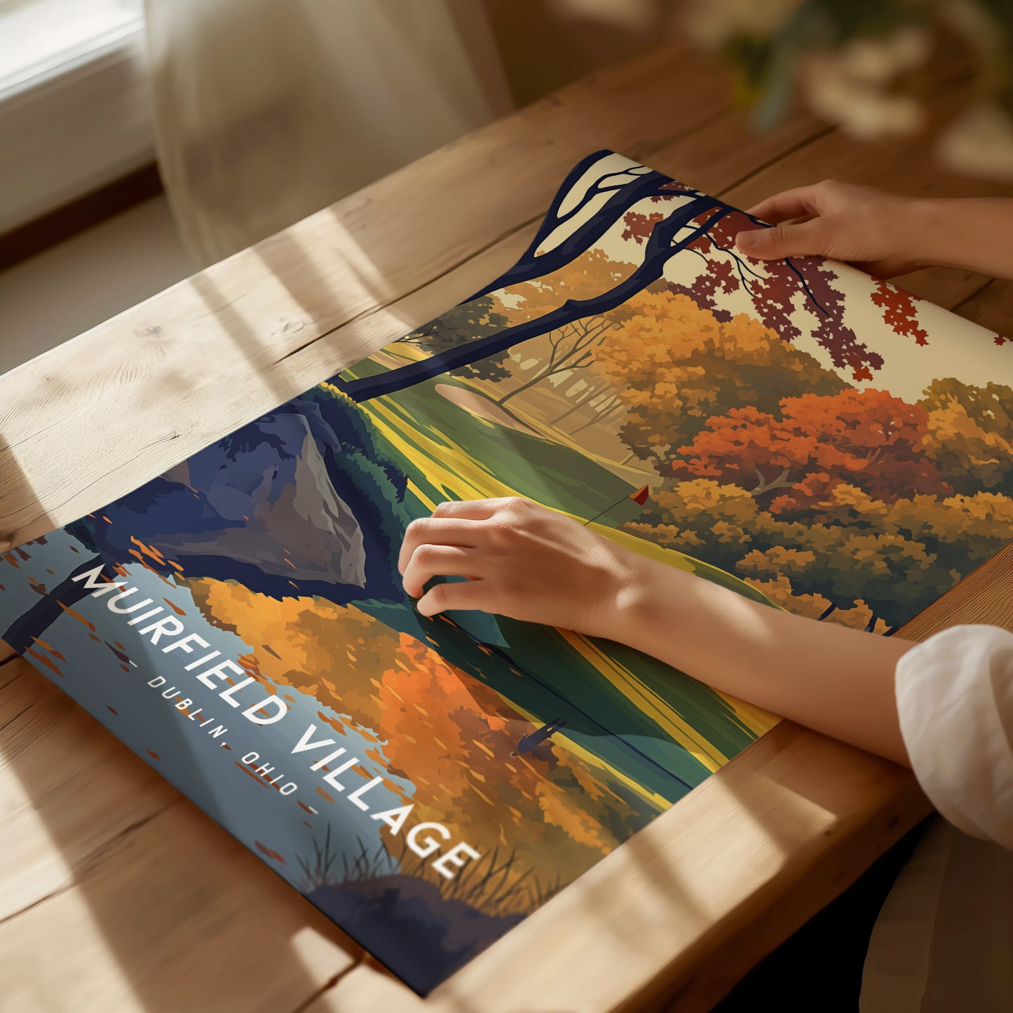 A person is arranging a colorful Muirfield Village poster on a wooden table, showcasing vibrant autumn trees and a golf course. Sunlight streams through a nearby window.