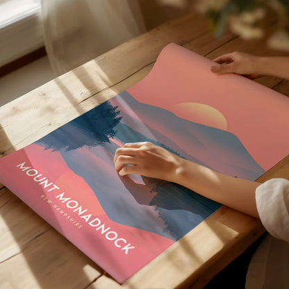 A person unrolls a colorful Mount Monadnock poster on a wooden table, featuring a stylized landscape with a pink sunset and trees. Sunlight casts gentle shadows across the scene.