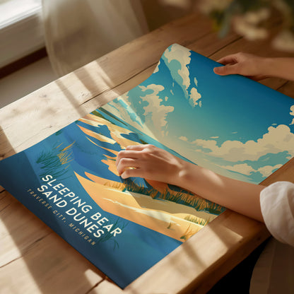 A person holds a Sleeping Bear Sand Dunes poster on a wooden table, featuring vibrant blue skies and golden sand dunes. Sunlight casts gentle shadows across the scene.