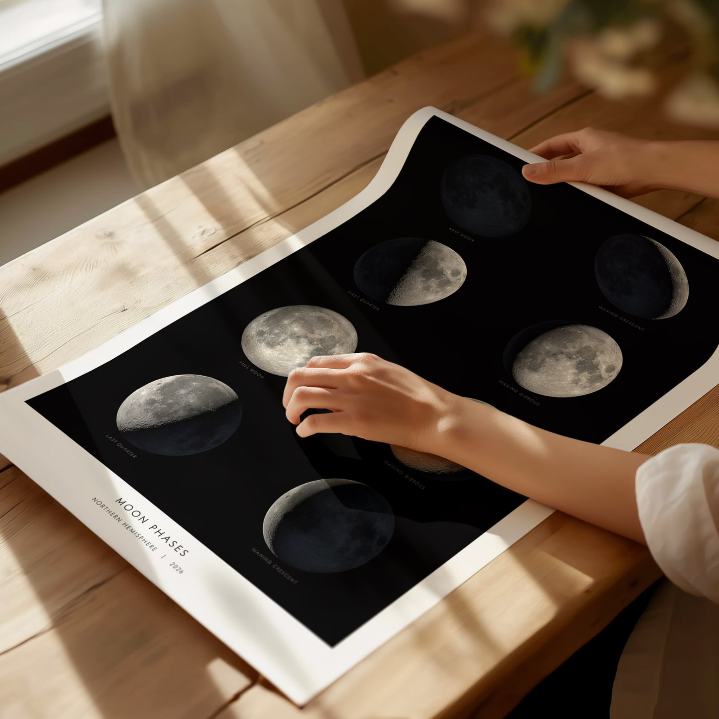 A person is gently touching a Moon Phases poster displayed on a wooden table. The poster features stunning images of the eight lunar phases, set against a dark background, highlighting the detailed textures of the moons surface.