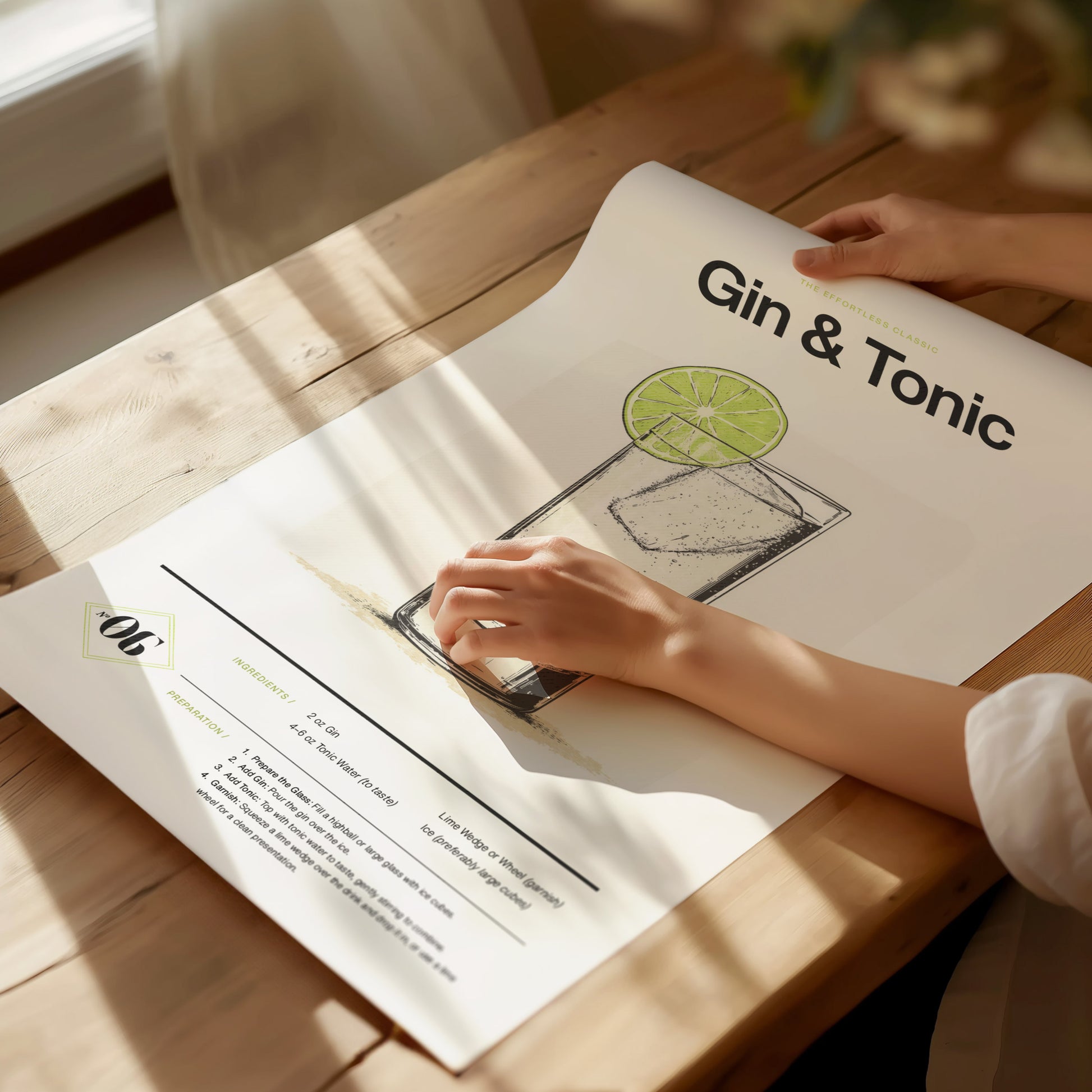 A person unrolls a Gin & Tonic poster on a wooden table, featuring an illustration of a cocktail with a lime slice. Sunlight casts soft shadows across the scene.