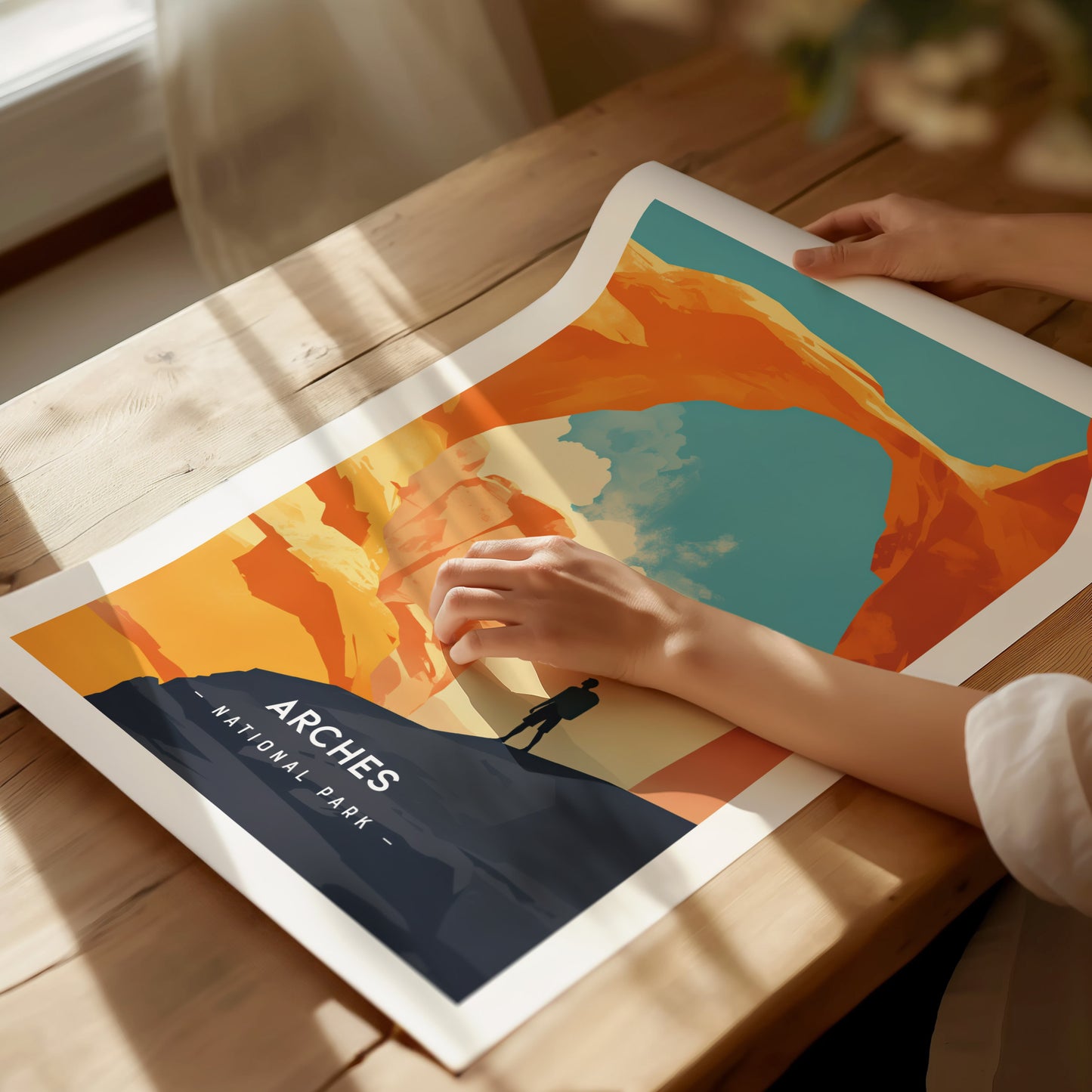A person examines a vibrant Arches National Park poster on a wooden table. The artwork features Delicate Arch against a bright blue sky, with sunlight streaming through a nearby window.