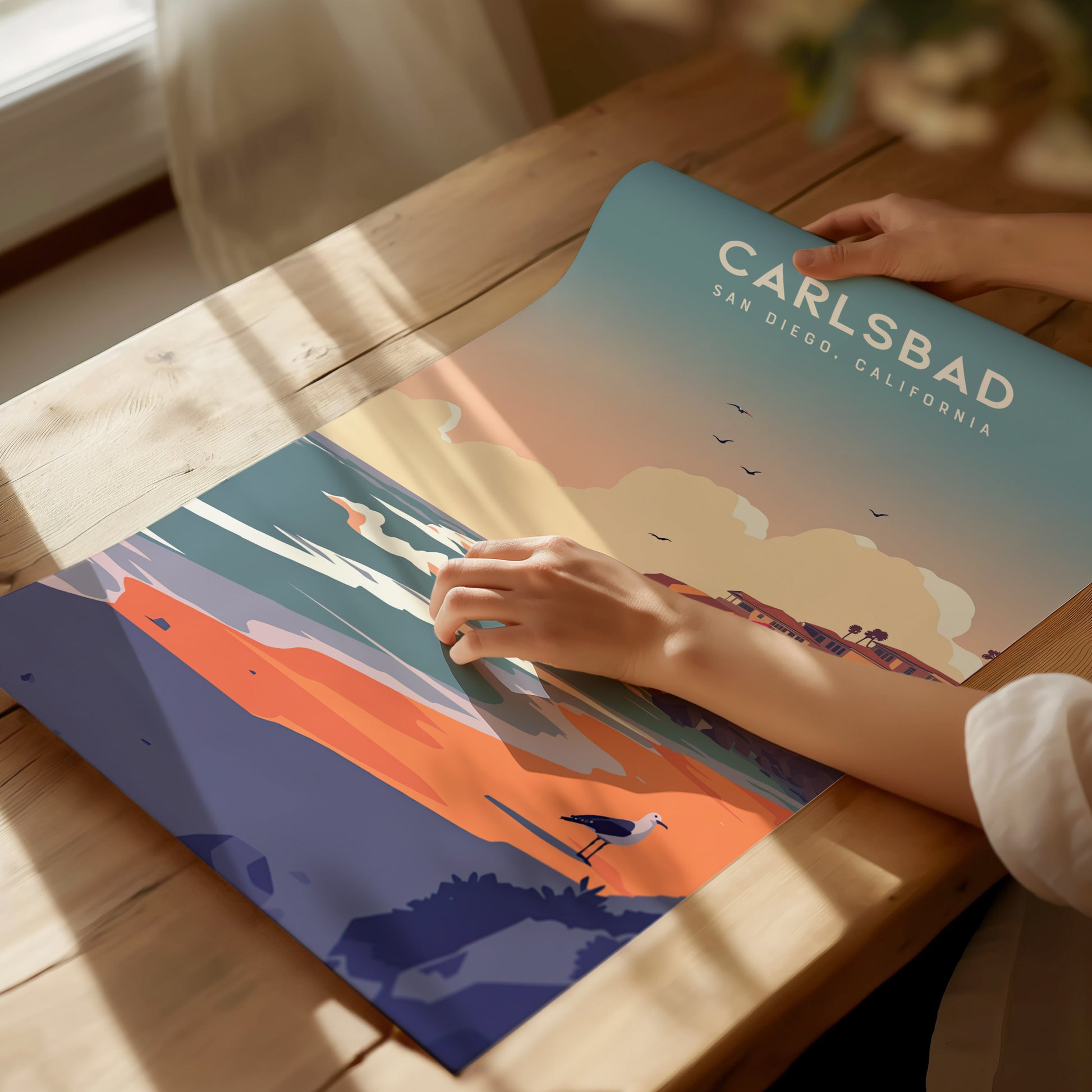 A person unrolls a colorful Carlsbad, San Diego poster on a wooden table, featuring vibrant coastal scenery with cliffs, beaches, and a seagull. Sunlight streams through a nearby window.