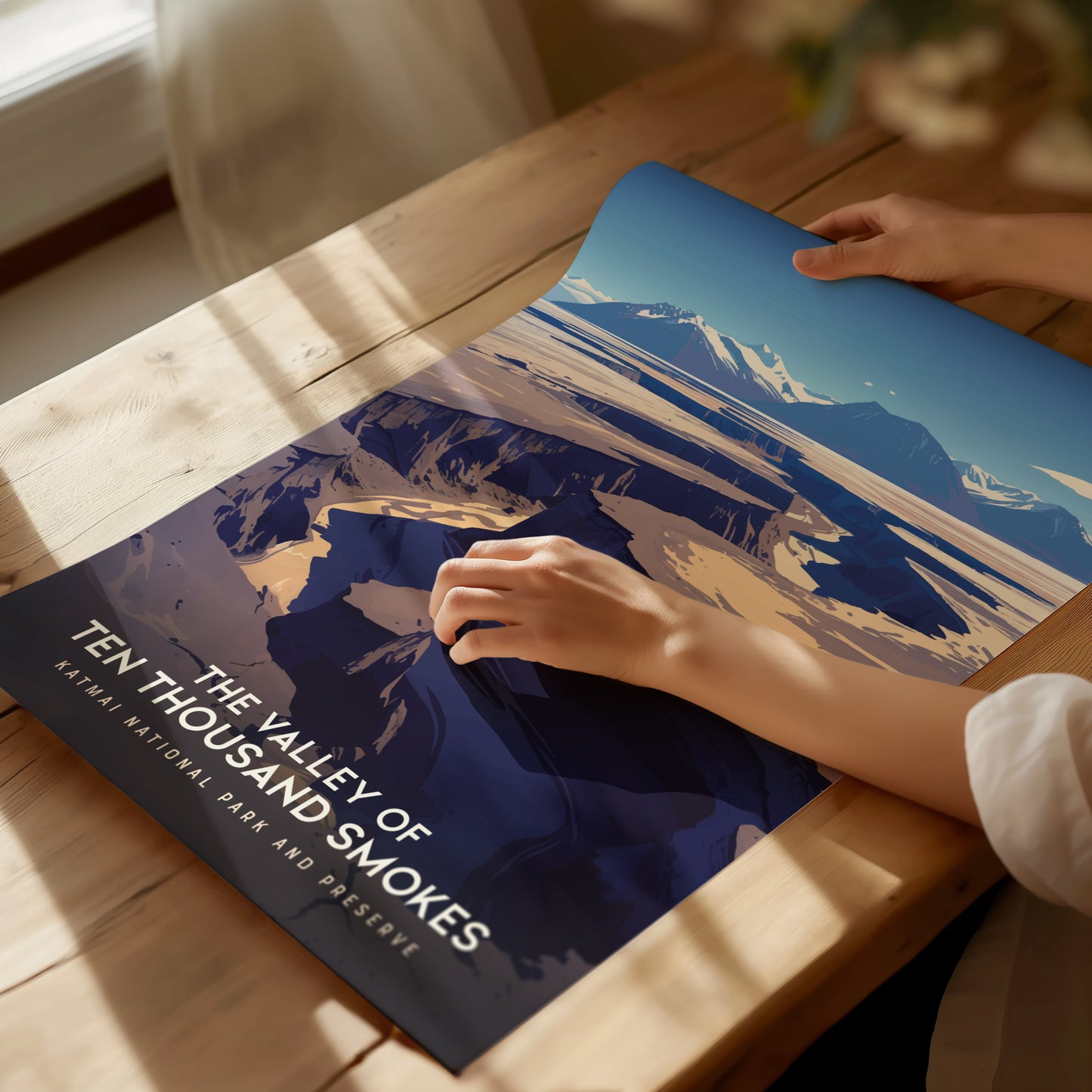 A person gently places a poster titled The Valley of Ten Thousand Smokes on a sunlit wooden table, showcasing a stunning volcanic landscape scene.