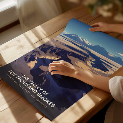 A person gently places a poster titled The Valley of Ten Thousand Smokes on a sunlit wooden table, showcasing a stunning volcanic landscape scene.
