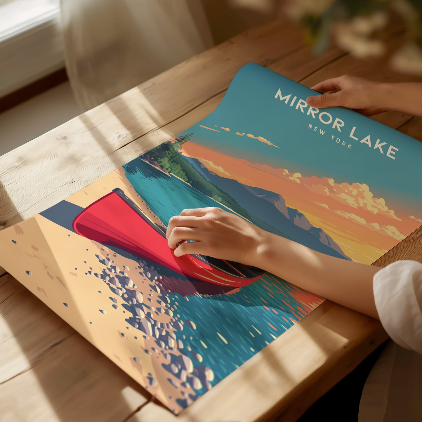 A person is holding a vibrant poster featuring Mirror Lake, New York, showcasing a red canoe on the water with colorful mountains and a sunset in the background. The scene conveys a serene and picturesque view of the Adirondacks.