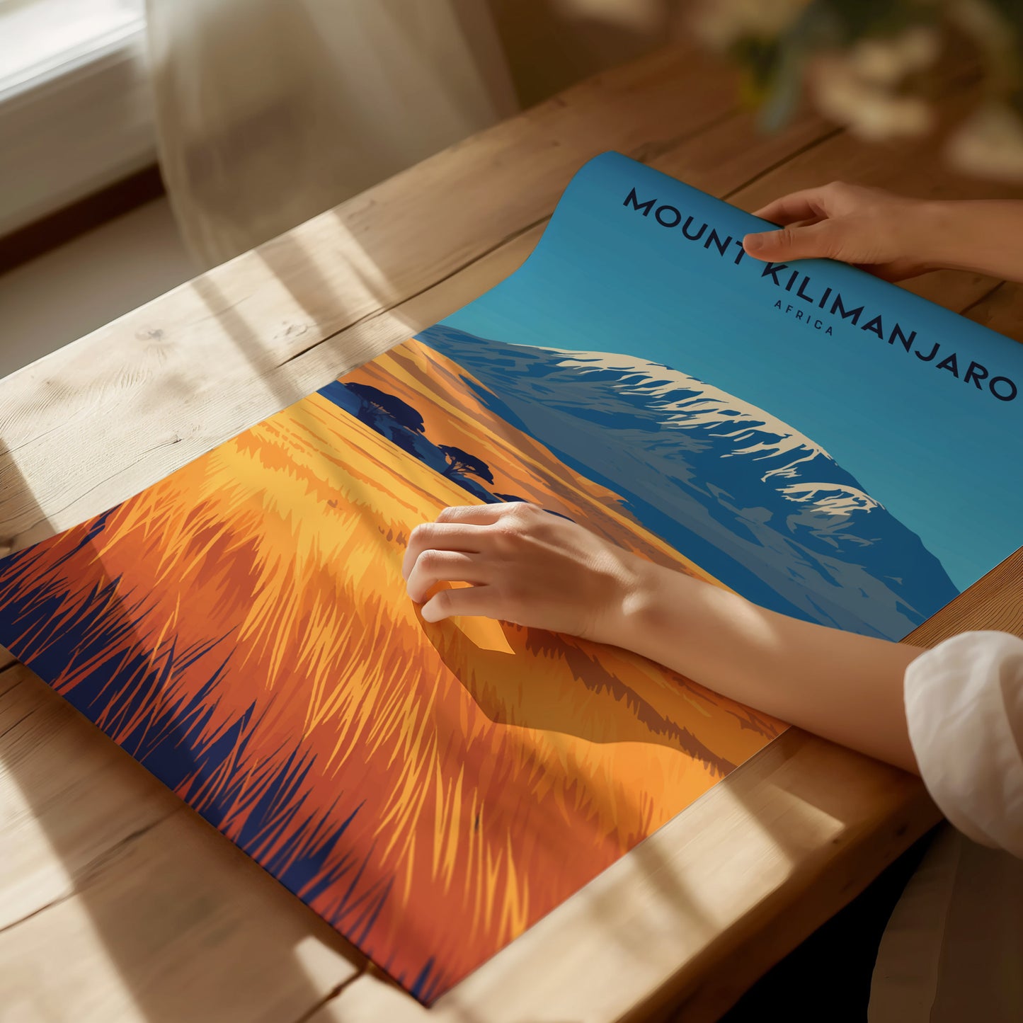 A person unrolls a colorful Mount Kilimanjaro poster on a wooden table, showcasing the mountains snow-capped peak and vibrant savanna landscape under a clear blue sky.