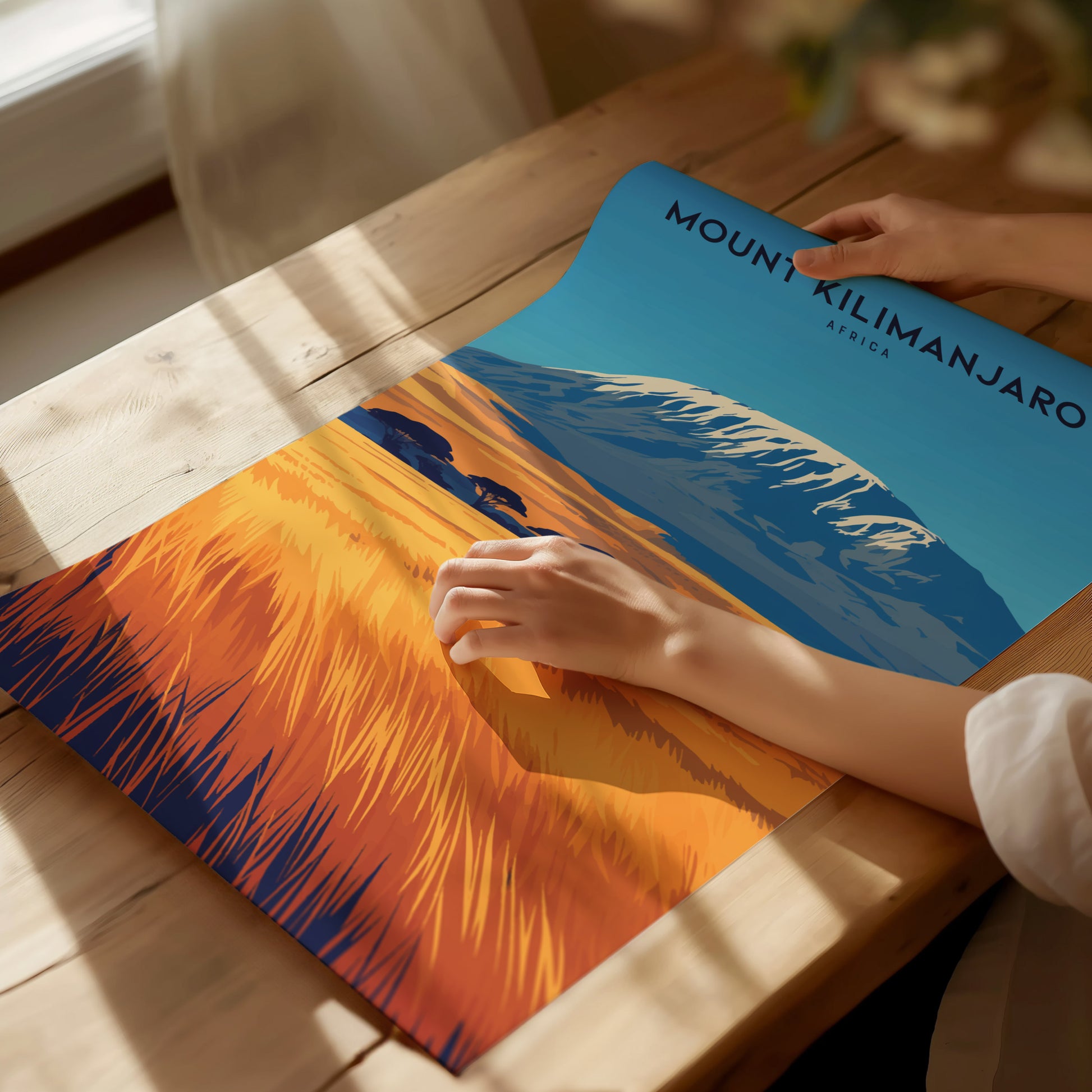A person unrolls a colorful Mount Kilimanjaro poster on a wooden table, showcasing the mountains snow-capped peak and vibrant savanna landscape under a clear blue sky.