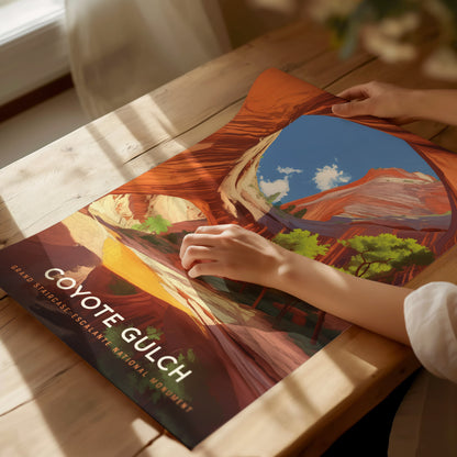 A person holds a colorful poster of Coyote Gulch, depicting vibrant canyon walls and a clear blue sky, on a sunlit wooden table. The text reads Coyote Gulch, Grand Staircase–Escalante.
