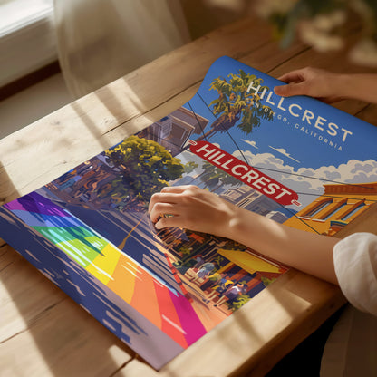 A person holds a colorful Hillcrest, San Diego poster featuring a vibrant rainbow crosswalk and the iconic Hillcrest sign, set on a wooden table in warm.