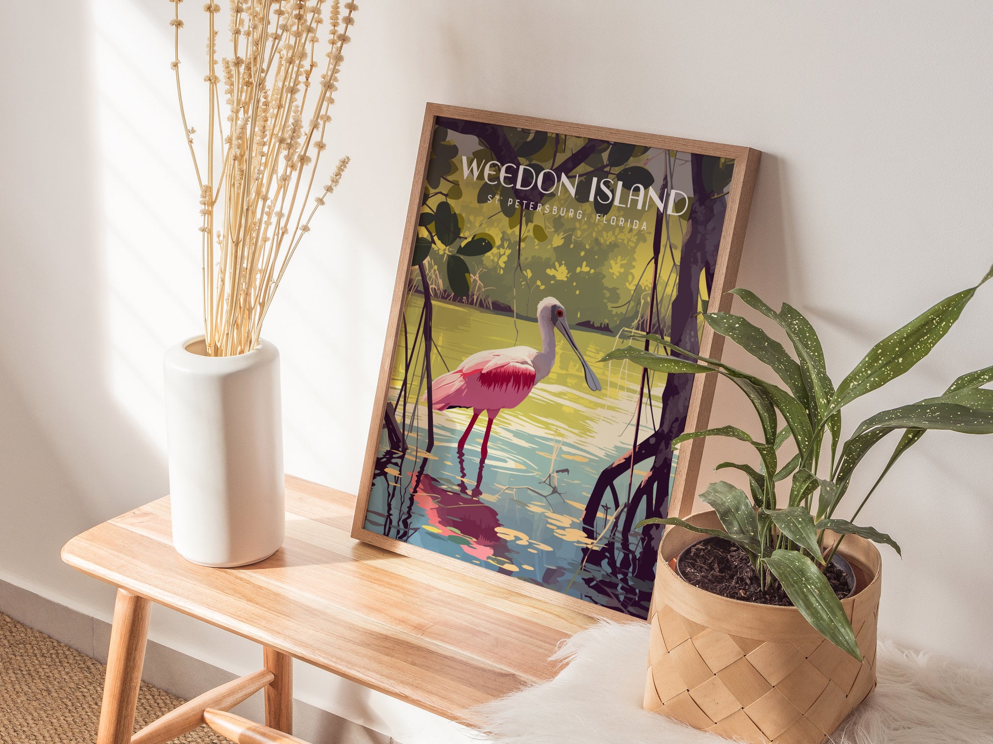 A framed poster depicting a pink Roseate Spoonbill wading in mangrove waters is placed on a wooden bench next to a white vase with dried flowers and a potted plant.