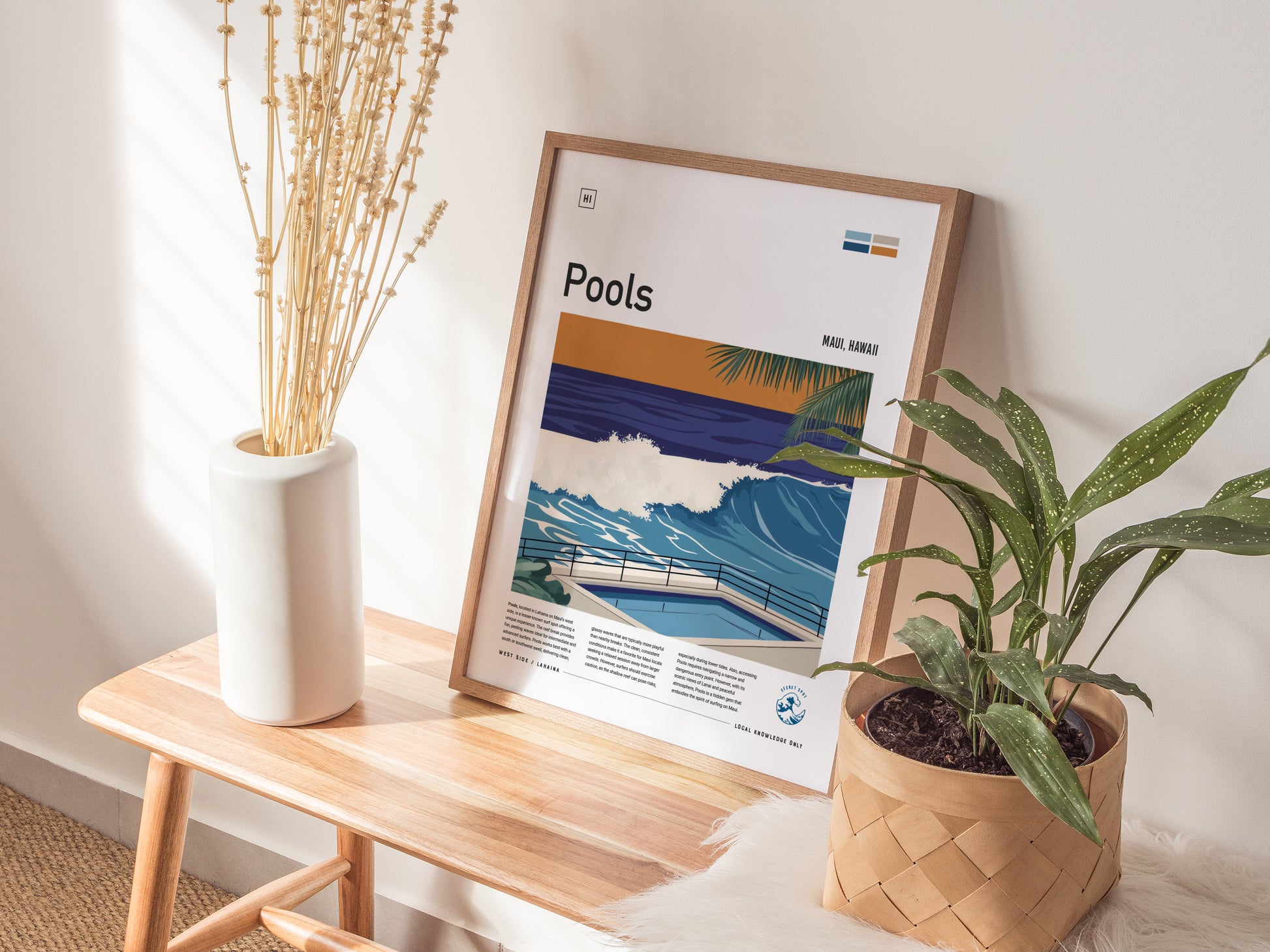 A framed surf art poster titled Pools featuring waves leans against a wall on a wooden bench, alongside a white vase with dried flowers and a potted plant.