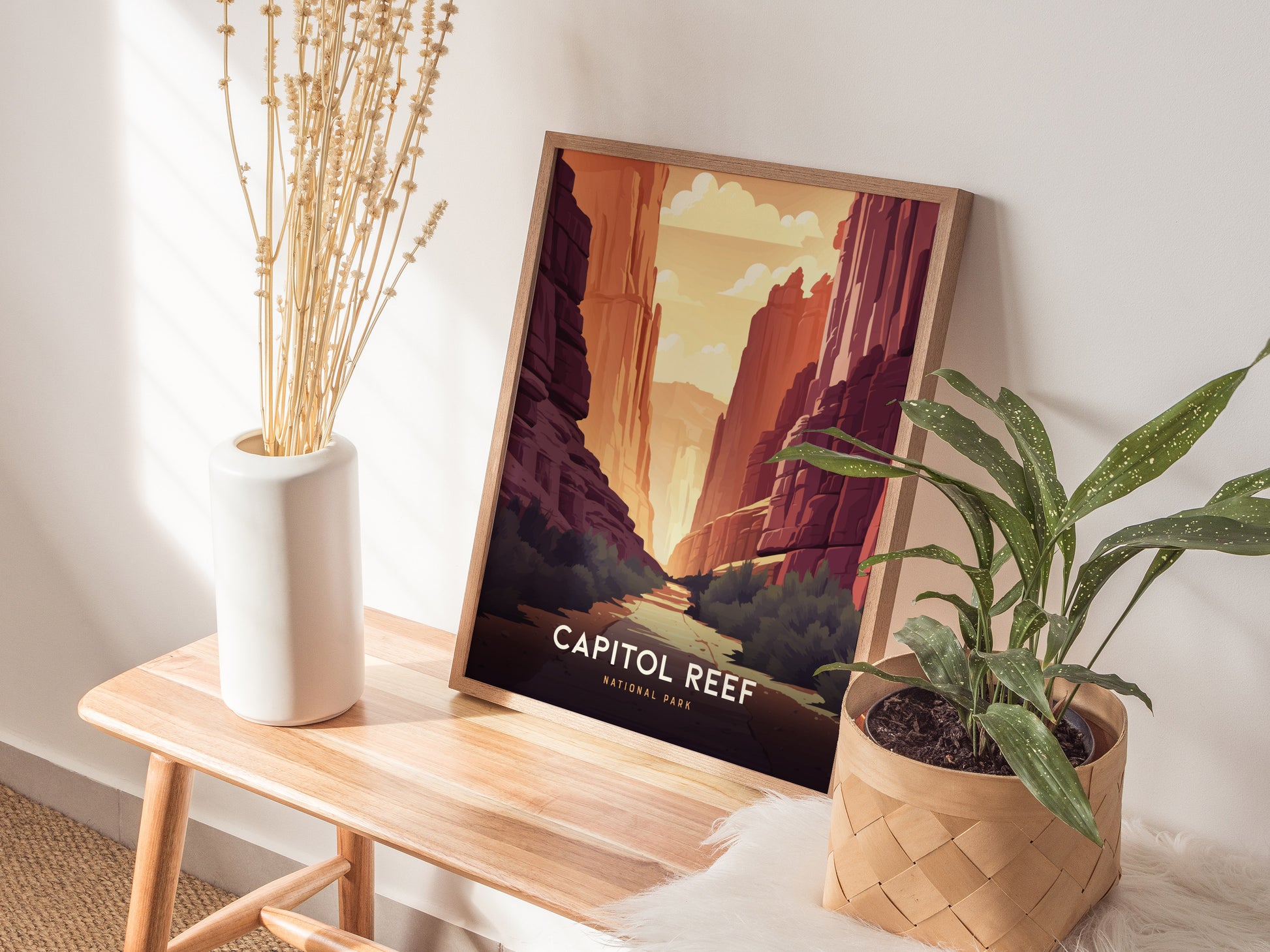 A framed Capitol Reef National Park poster rests on a wooden bench between a vase with dried stems and a leafy potted plant, creating a cozy, natural display.