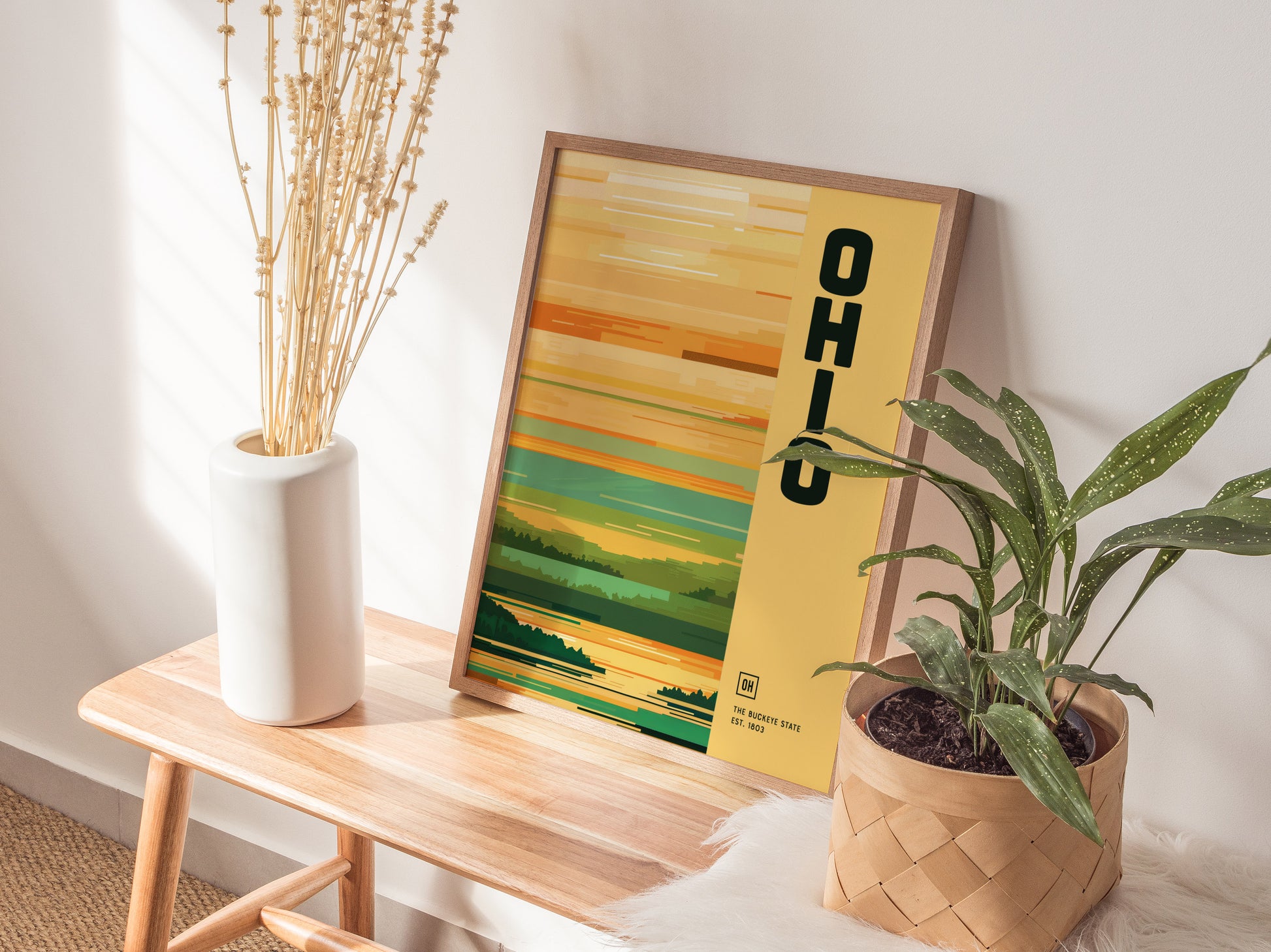 A minimalist Ohio poster with green and yellow tones rests on a wooden bench next to a white vase with dried flowers and a potted plant.