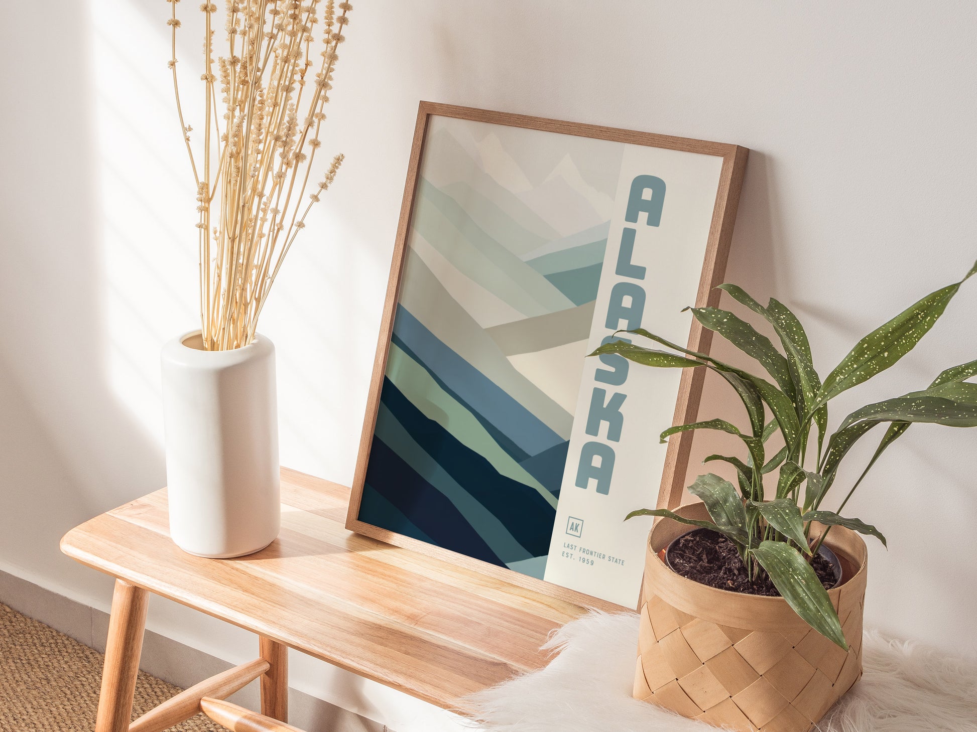 A minimalist Alaska-themed poster with abstract mountains in blues and grays leans against a white wall. It sits on a wooden bench next to a white vase and a potted.