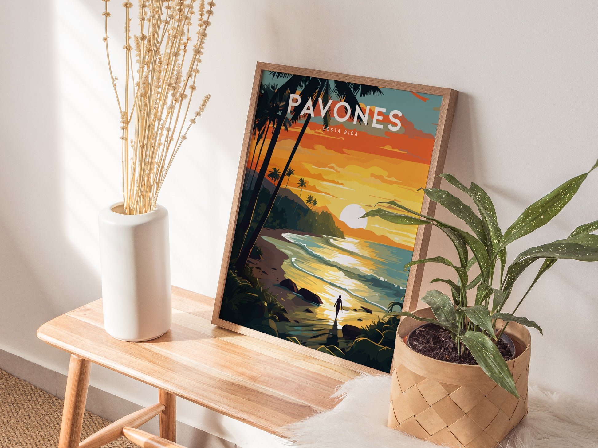 A framed surf poster with a vibrant sunset beach scene rests on a wooden bench beside a white vase with tall dried stems and a potted plant.