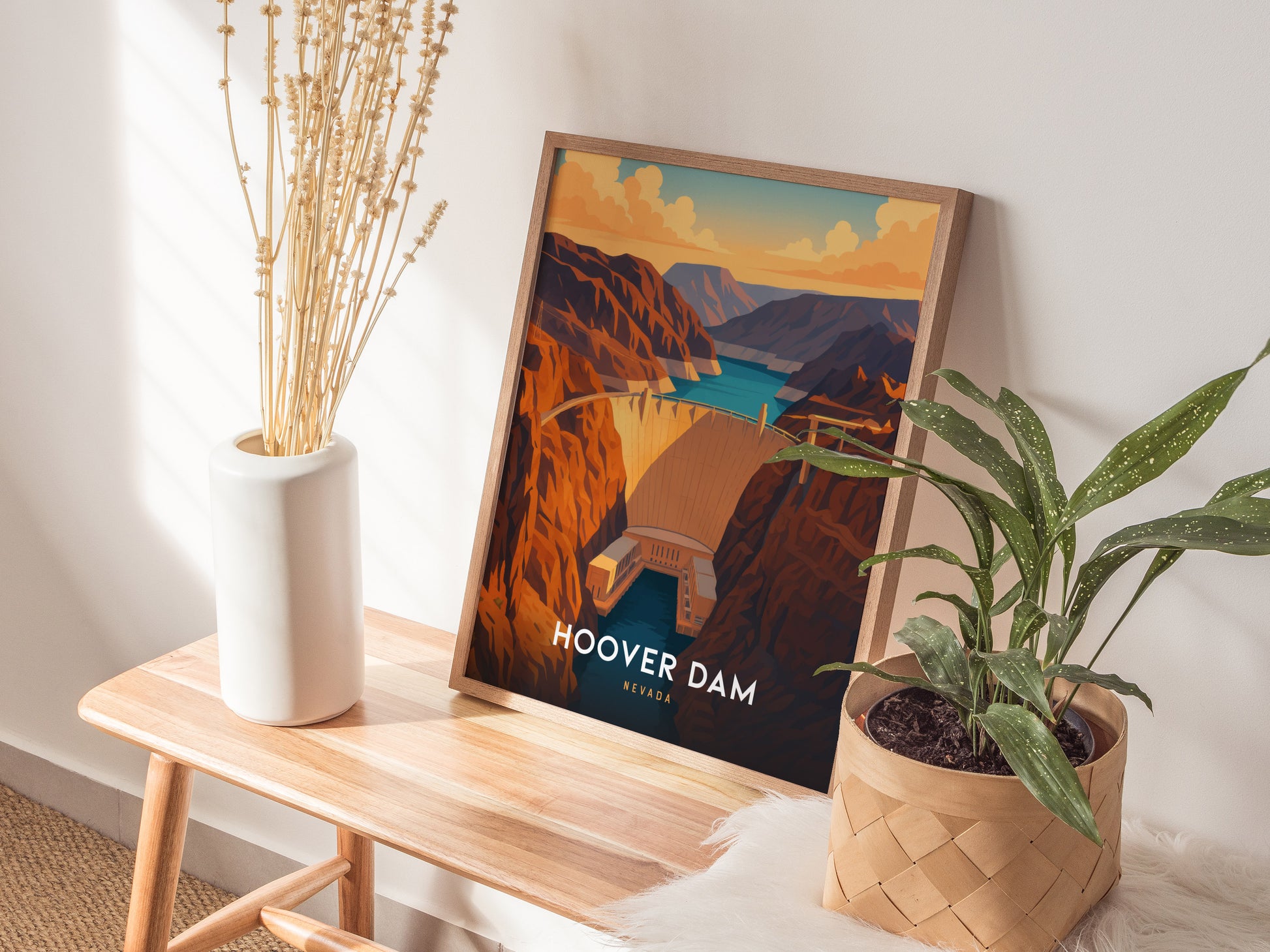 A framed illustration of the Hoover Dam rests on a wooden bench, alongside a white vase with dried plants and a woven basket containing a green plant. Sunlight casts soft shadows on the wall, creating a warm, inviting atmosphere.