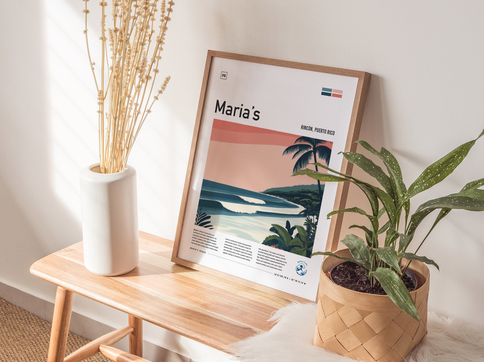 A framed poster titled Marias showcasing a tropical surf scene rests on a wooden bench, accompanied by a white vase with dried branches and a potted green plant.