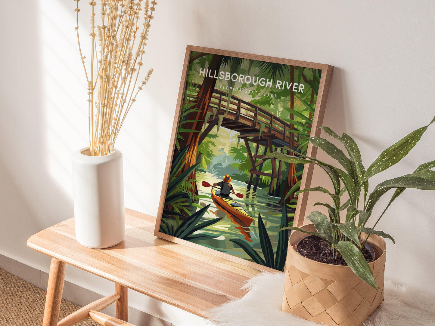 A framed Hillsborough River poster depicting a kayaker under a wooden bridge is displayed on a wooden bench, accompanied by a vase with dried flowers and a potted plant.