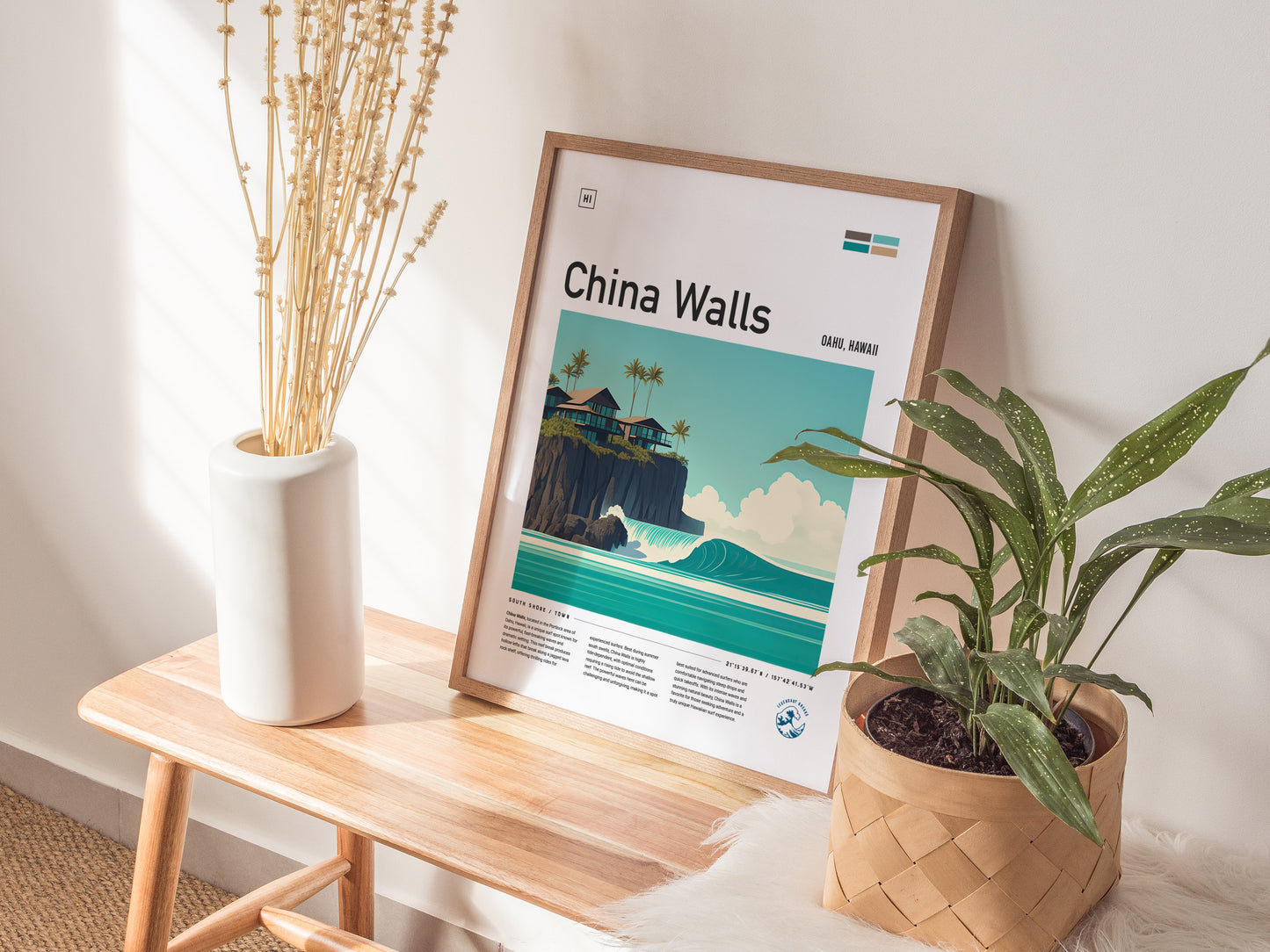 A framed poster titled China Walls depicting a coastal scene with waves and cliffs, rests on a wooden bench beside a vase with dried flowers and a potted plant.
