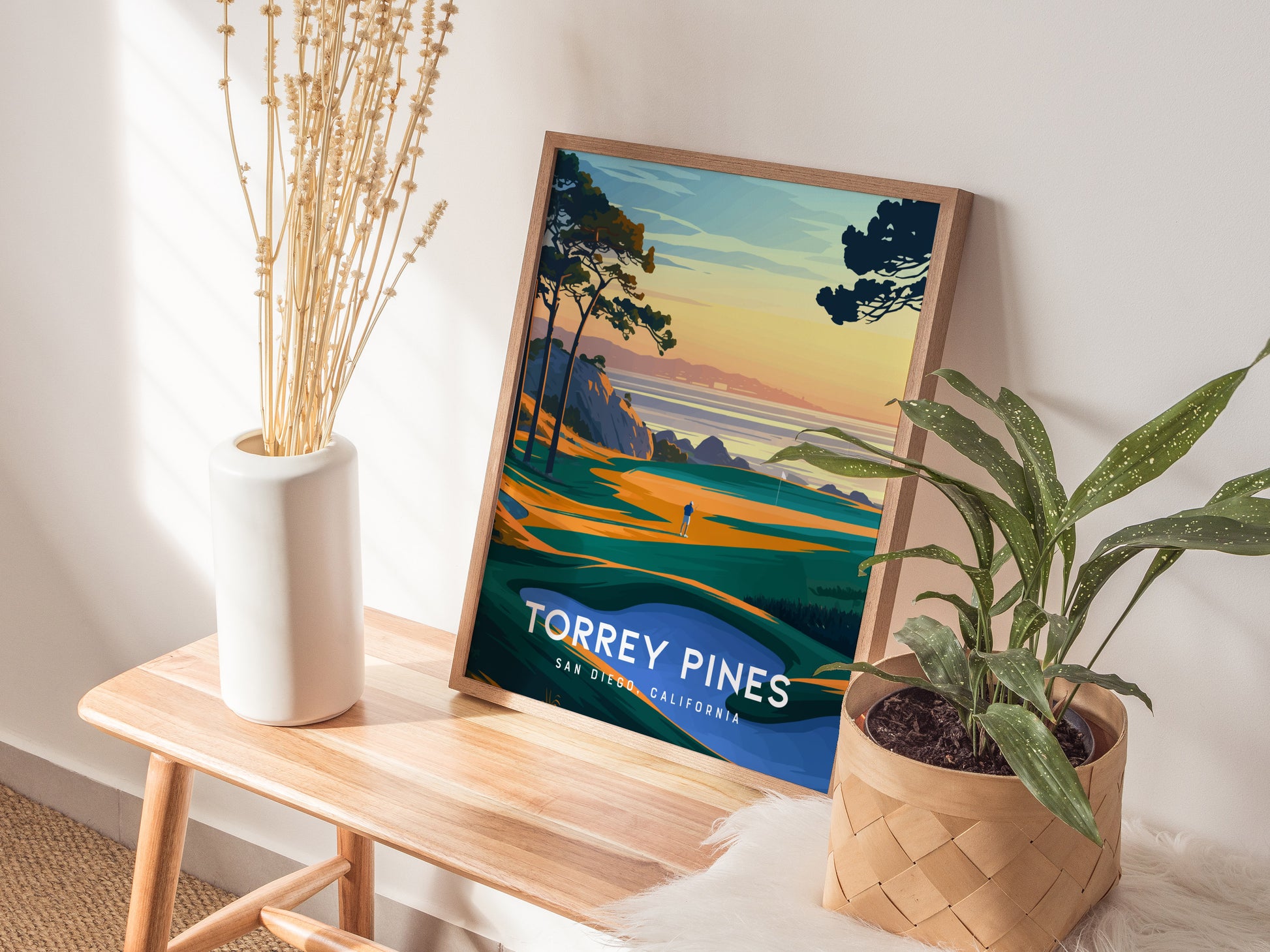 A framed Torrey Pines poster depicting a scenic golf course rests on a wooden bench, surrounded by a white vase with dried flowers and a green potted plant.