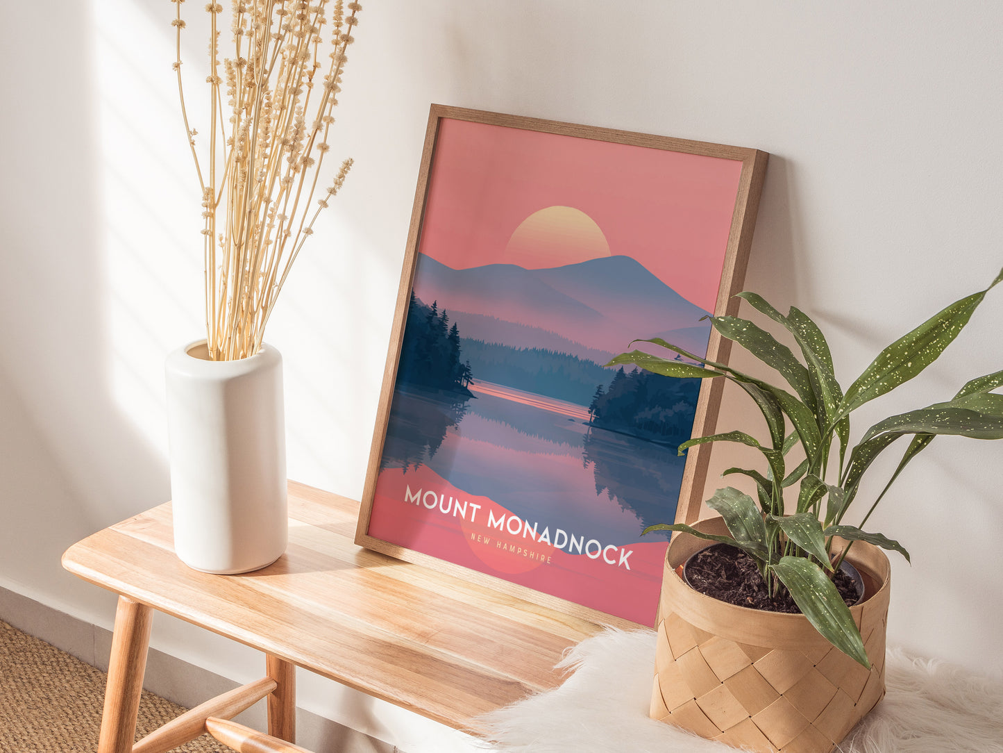 A Mount Monadnock poster with a pink sunset landscape leans against a wall on a wooden table, accompanied by a white vase with dried flowers and a potted plant.