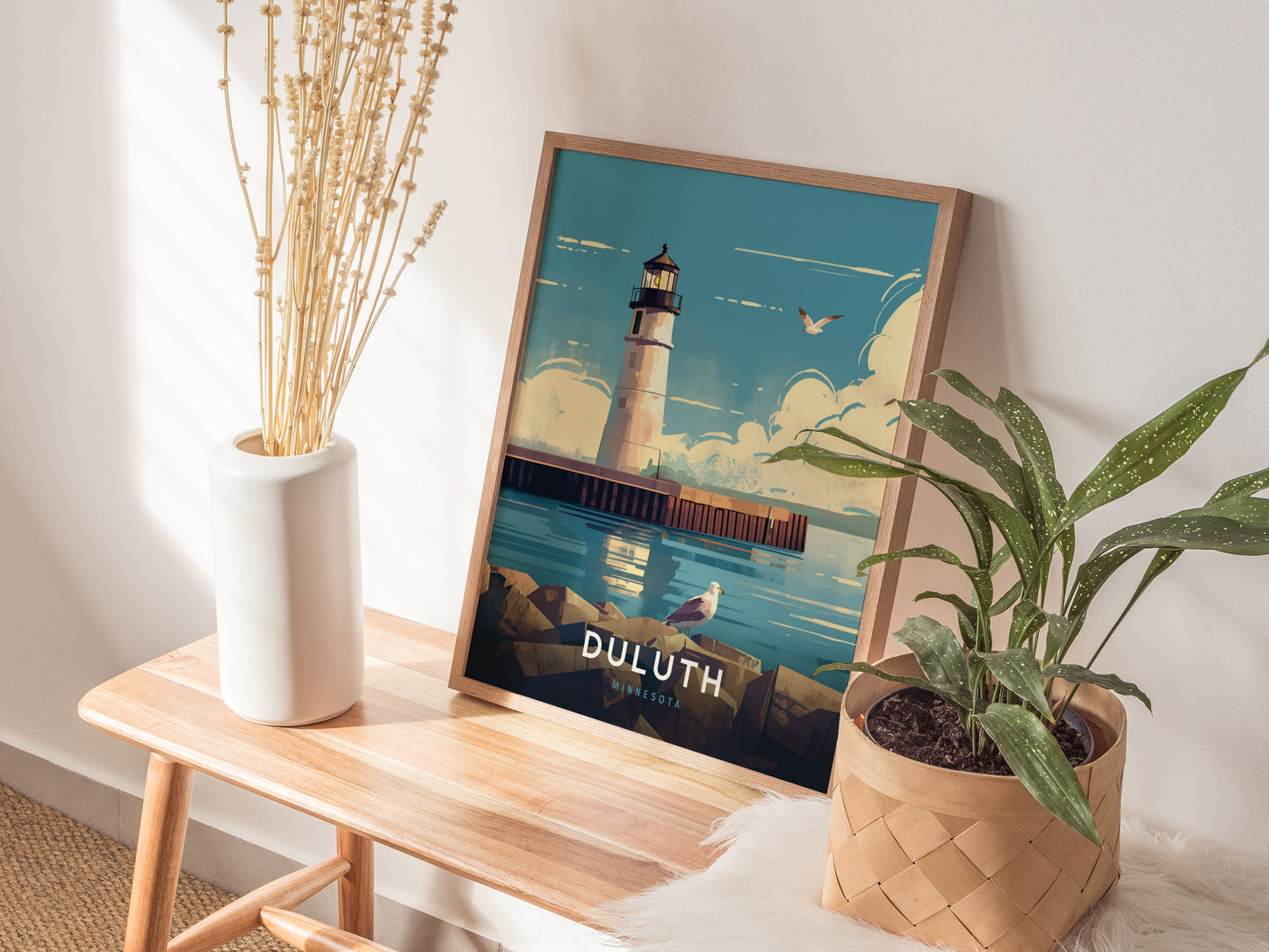 A framed poster of the Duluth Harbor North Pier Lighthouse is displayed on a wooden bench, surrounded by a white vase with dried flowers and a potted green plant.