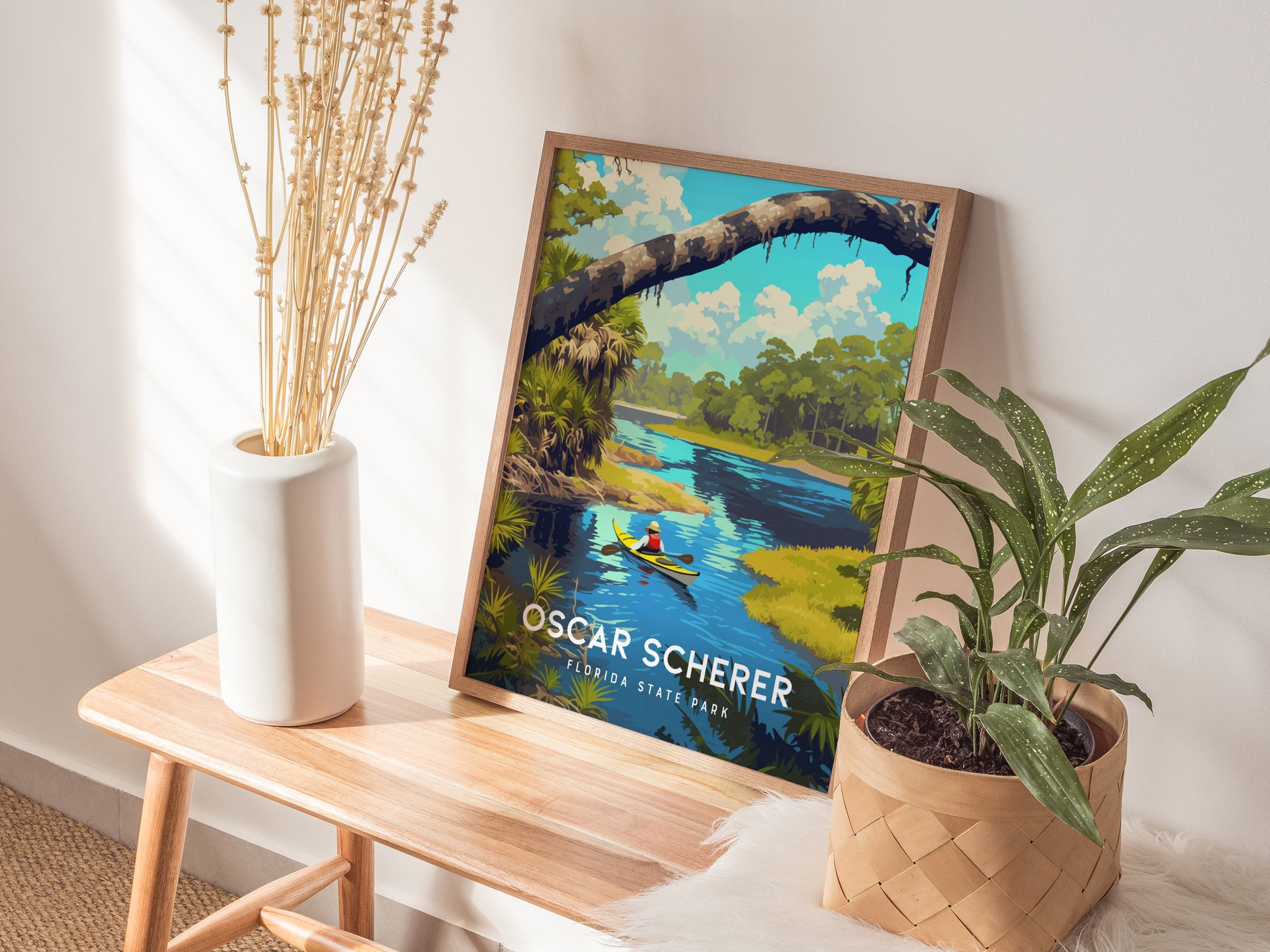 A framed Oscar Scherer State Park poster rests on a wooden bench alongside a white vase with dried flowers and a potted plant, creating a serene, natural decor setting.