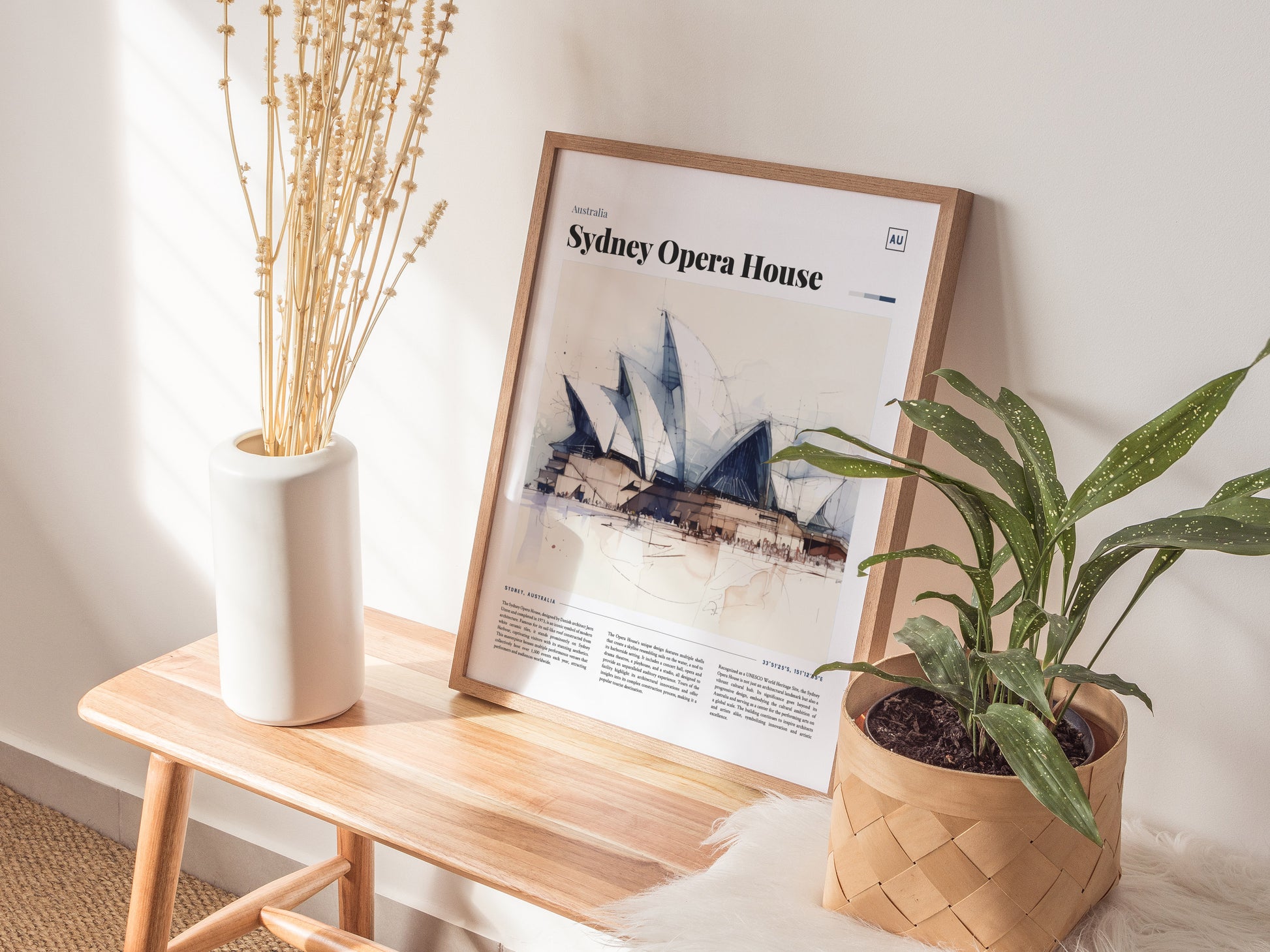 A framed Sydney Opera House poster sits on a wooden bench between a white vase with dried flowers and a potted green plant. Sunlight streams through, casting soft shadows.